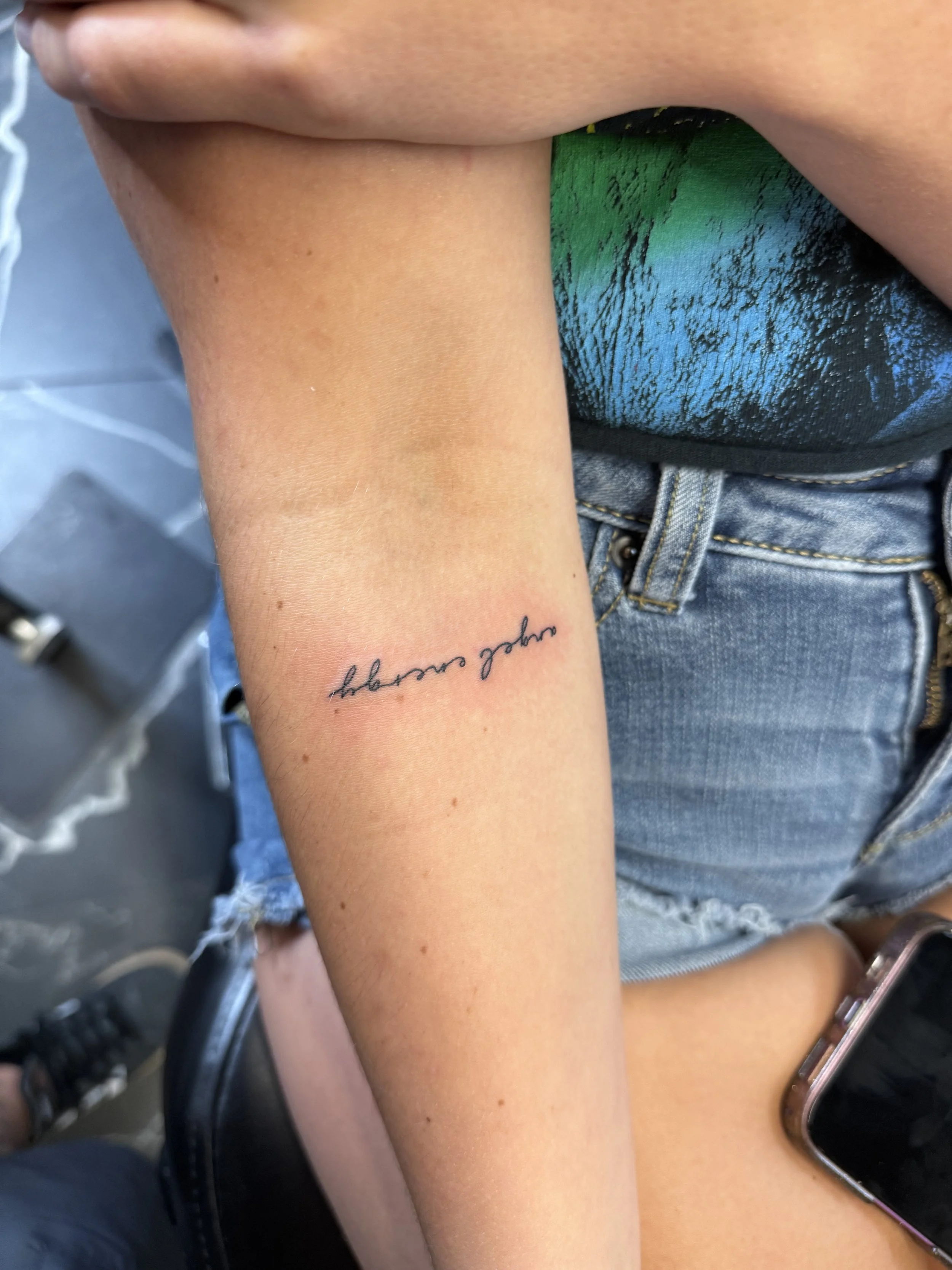 Close-up of a person's arm with a handwritten tattoo that says 'dream summer' in cursive, with part of a rainbow-colored shirt and denim shorts visible.