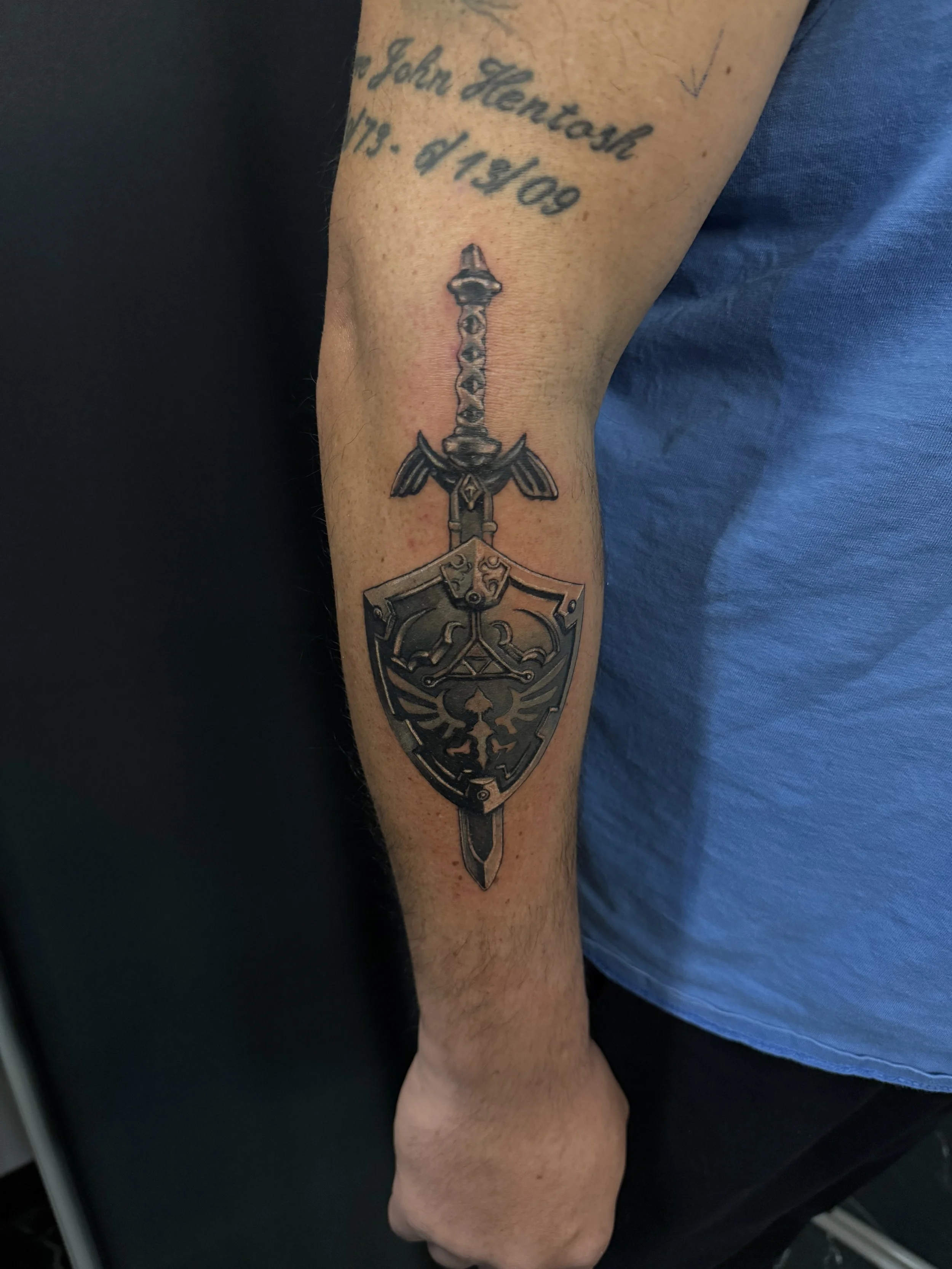 Tattoo of a sword with a shield on the inner forearm, with text above it that reads 'John Hentosh 1979 - 4/19/09'.