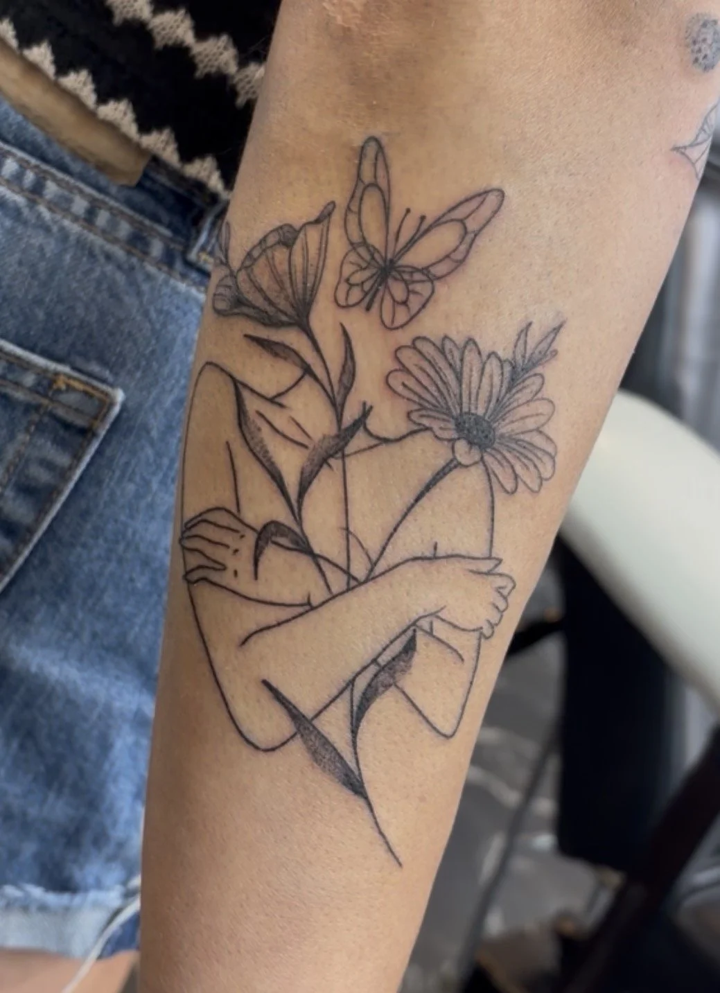 Black outline tattoo of a hand holding a daisy flower, with butterflies and additional flowers around it.
