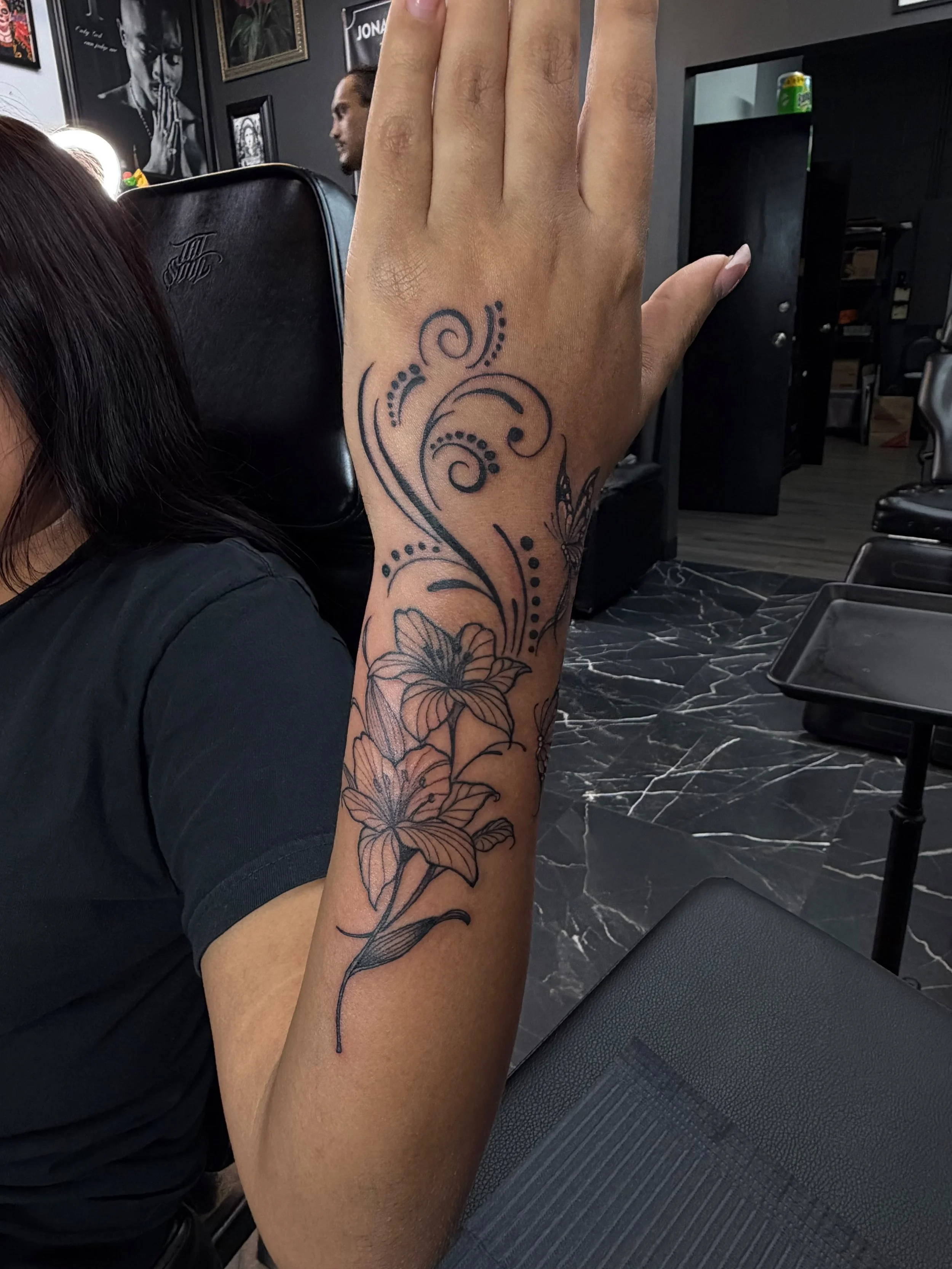 Close-up of a person's arm with a floral tattoo, raised in a tattoo studio with black walls, framed artwork, and a person sitting in the background.