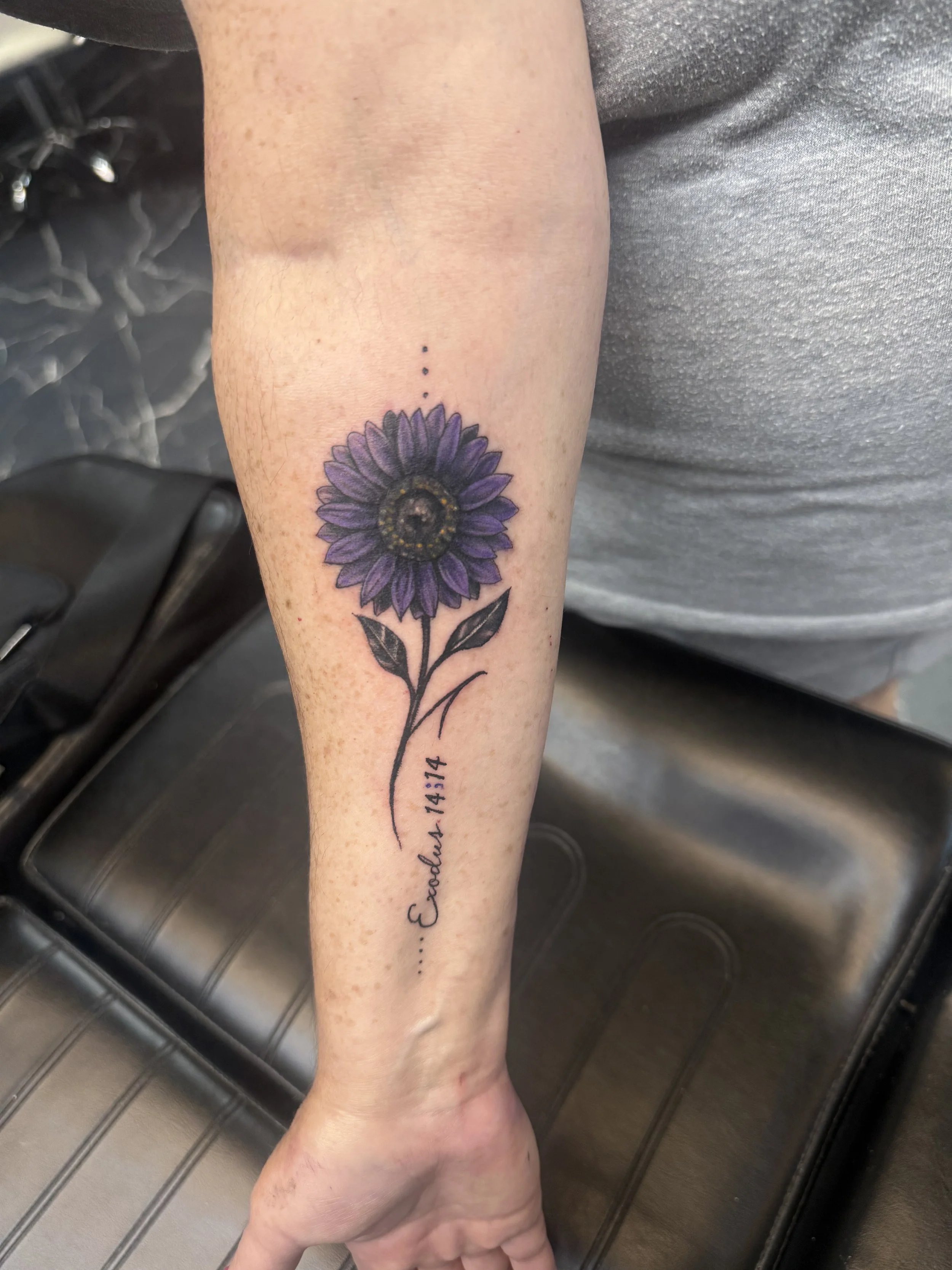 A person's forearm with a purple flower tattoo and black ink writing, resting on a metallic surface.