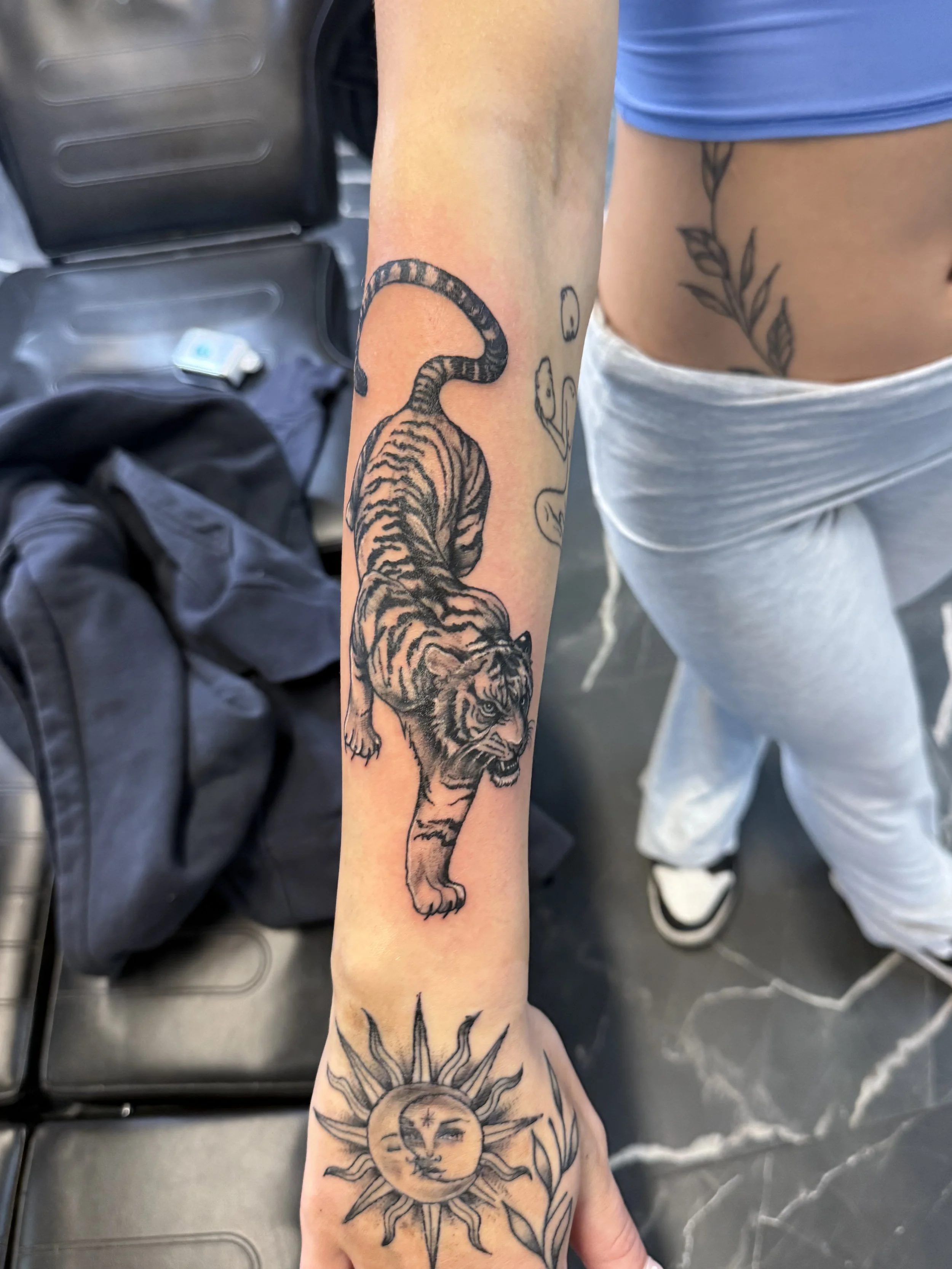Inner forearm tattoo of a tiger with detailed stripes and face, and a sun with a face, sun rays, and a moon in the background.