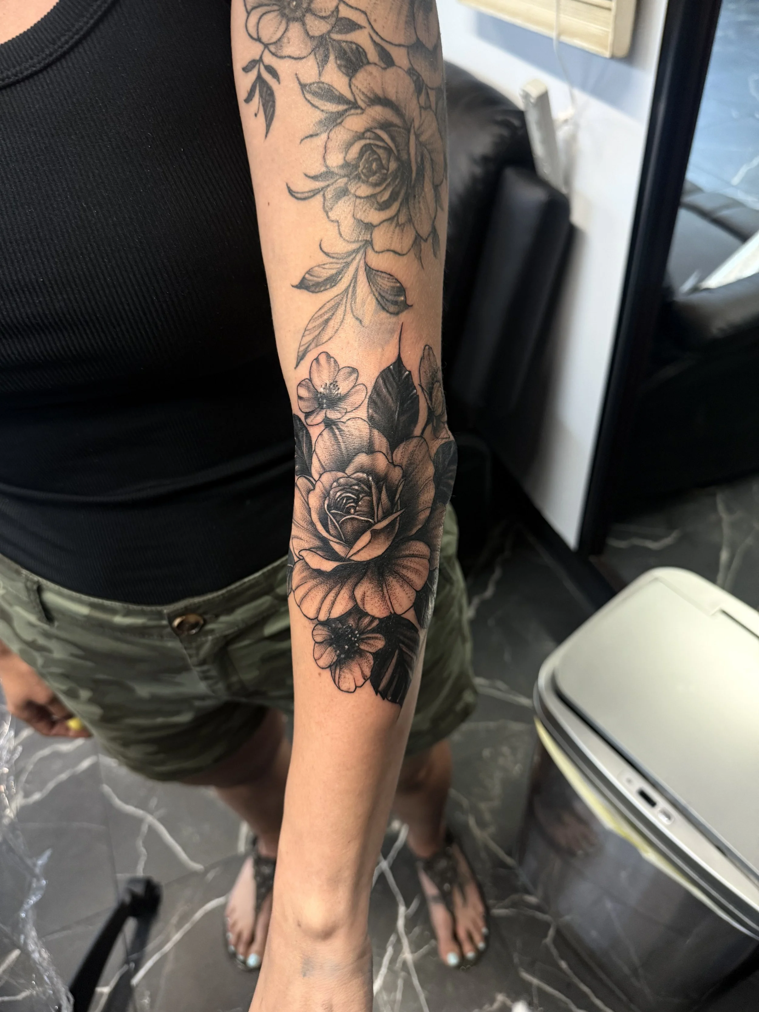 A person's arm with a black and gray floral tattoo featuring large roses, leaves, and smaller flowers. The person is wearing a black tank top and camouflage shorts, standing on a black and white marble floor.