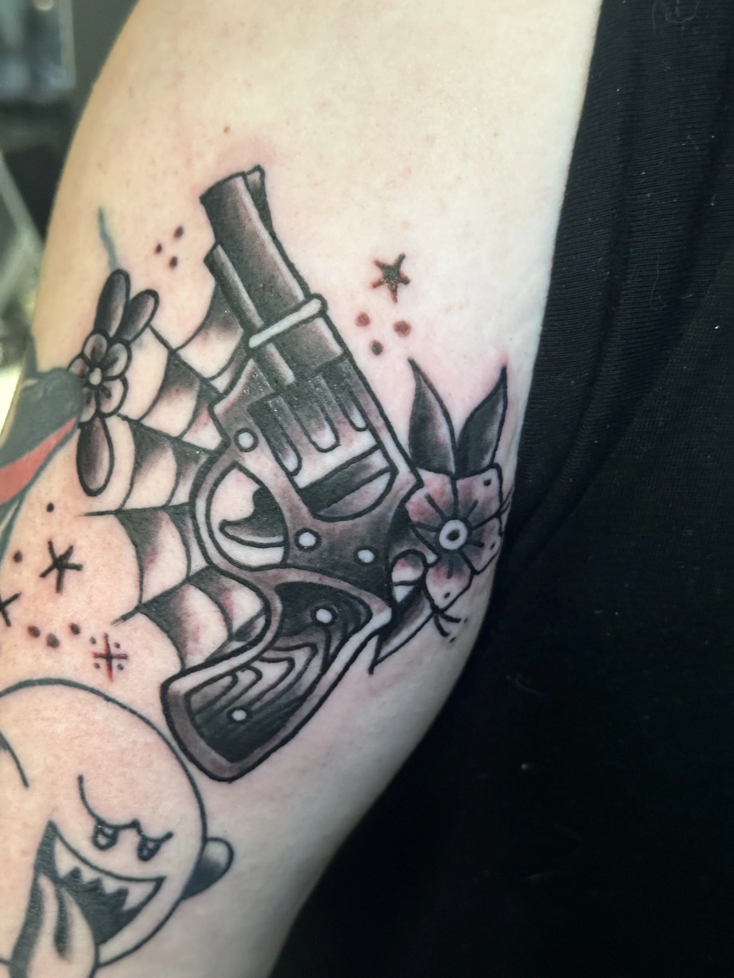 Tattoo of a cartoon-style handgun with a bow, flowers, stars, and dots on a person's arm.