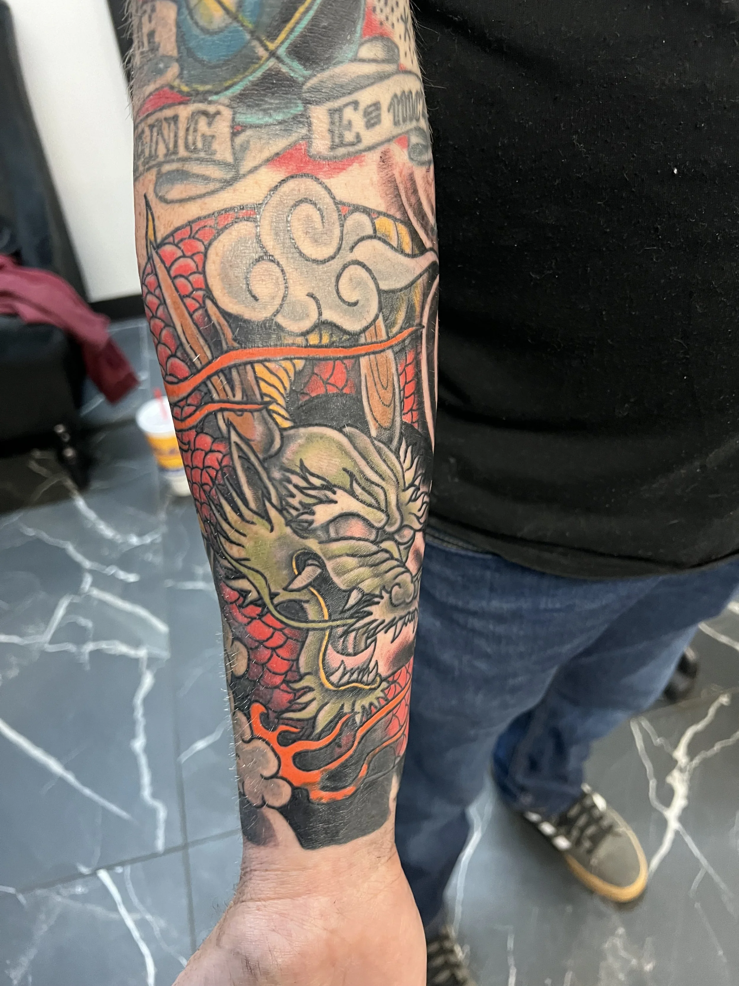 A person's tattooed arm showing a colorful tattoo of a lion's face with red and black scales, and a blue and red banner with text.
