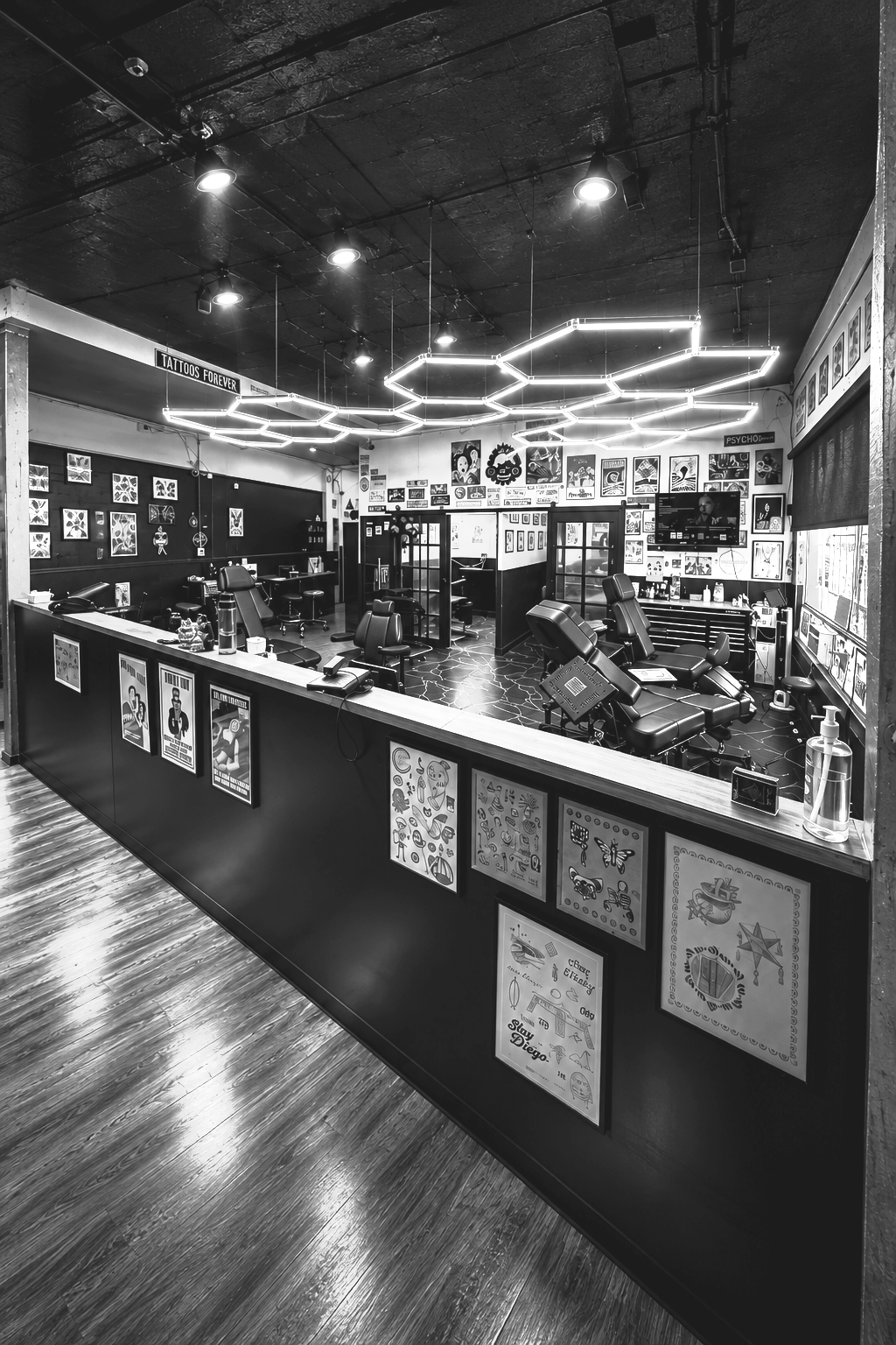 Black and white photo of a tattoo shop interior with various tattoo chairs, artwork on the walls, bright geometric ceiling lights, and tattoo station counters with design posters.