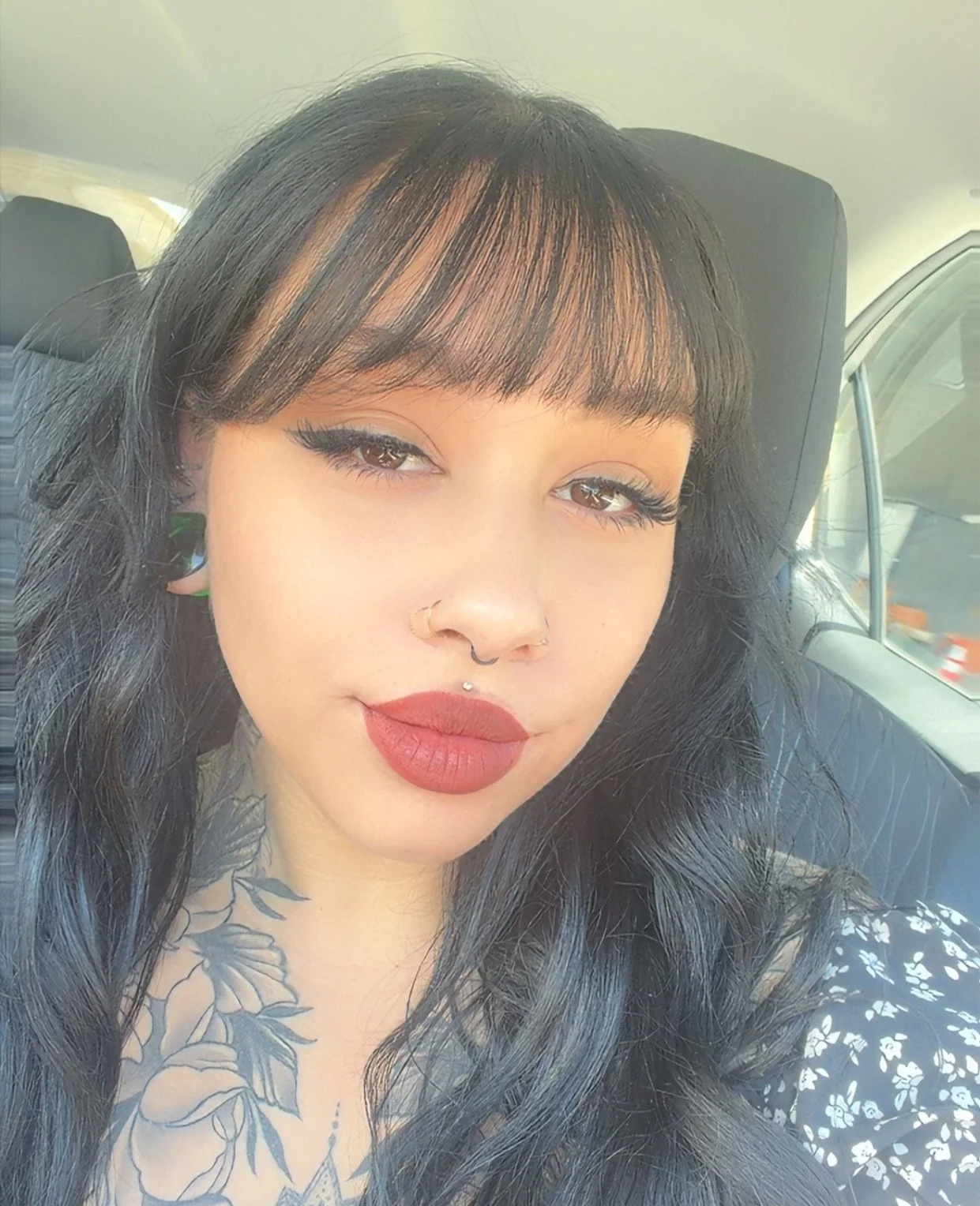 Close-up of a young woman with black hair and bangs, wearing makeup with dark lipstick and eyeliner, sitting in a car with a tattoo on her chest visible.