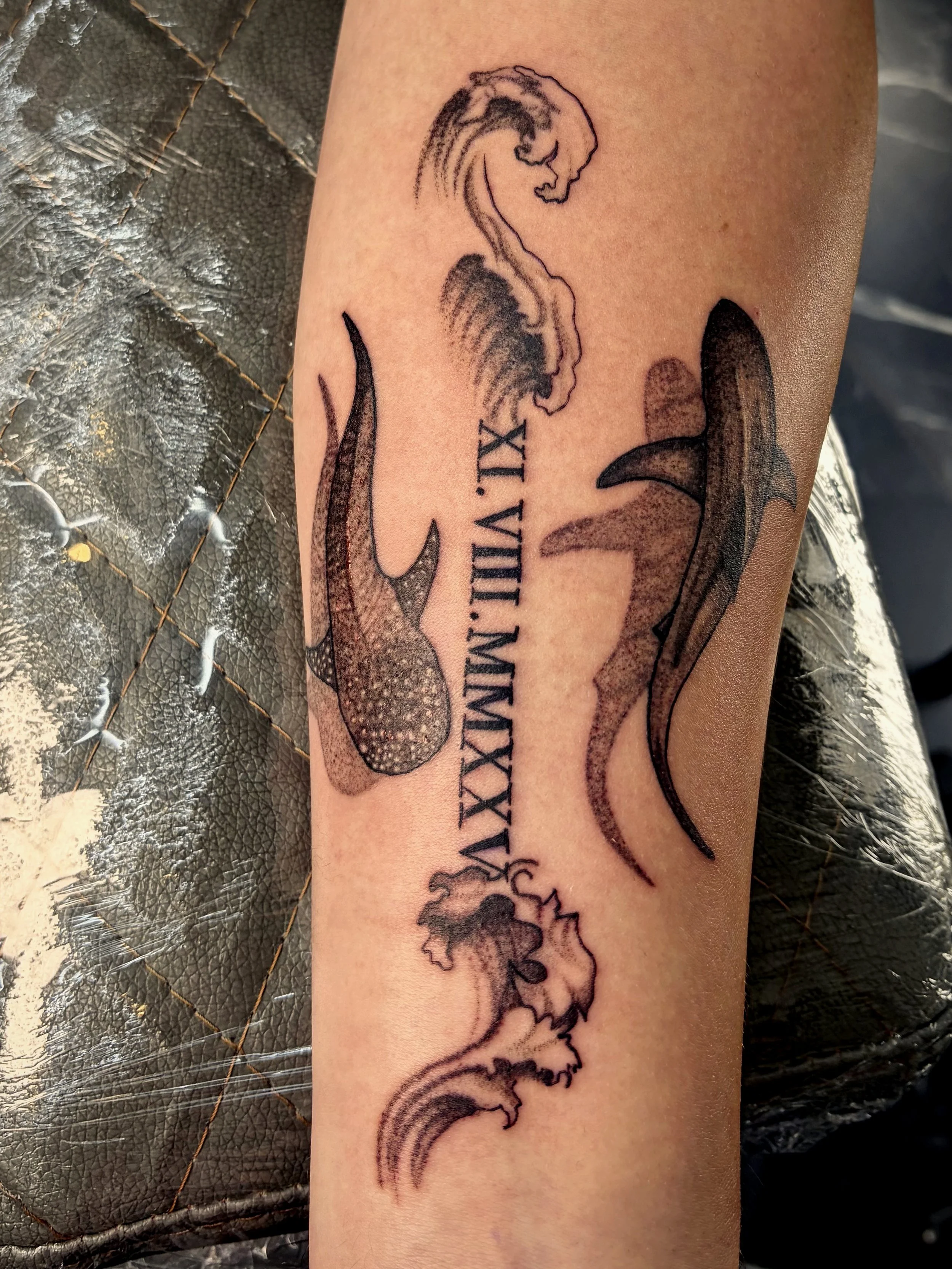 A tattoo on a person's forearm depicting a stylized dragon or mythical creature intertwined with a rainbow design. The tattoo also includes Roman numerals and some decorative elements.