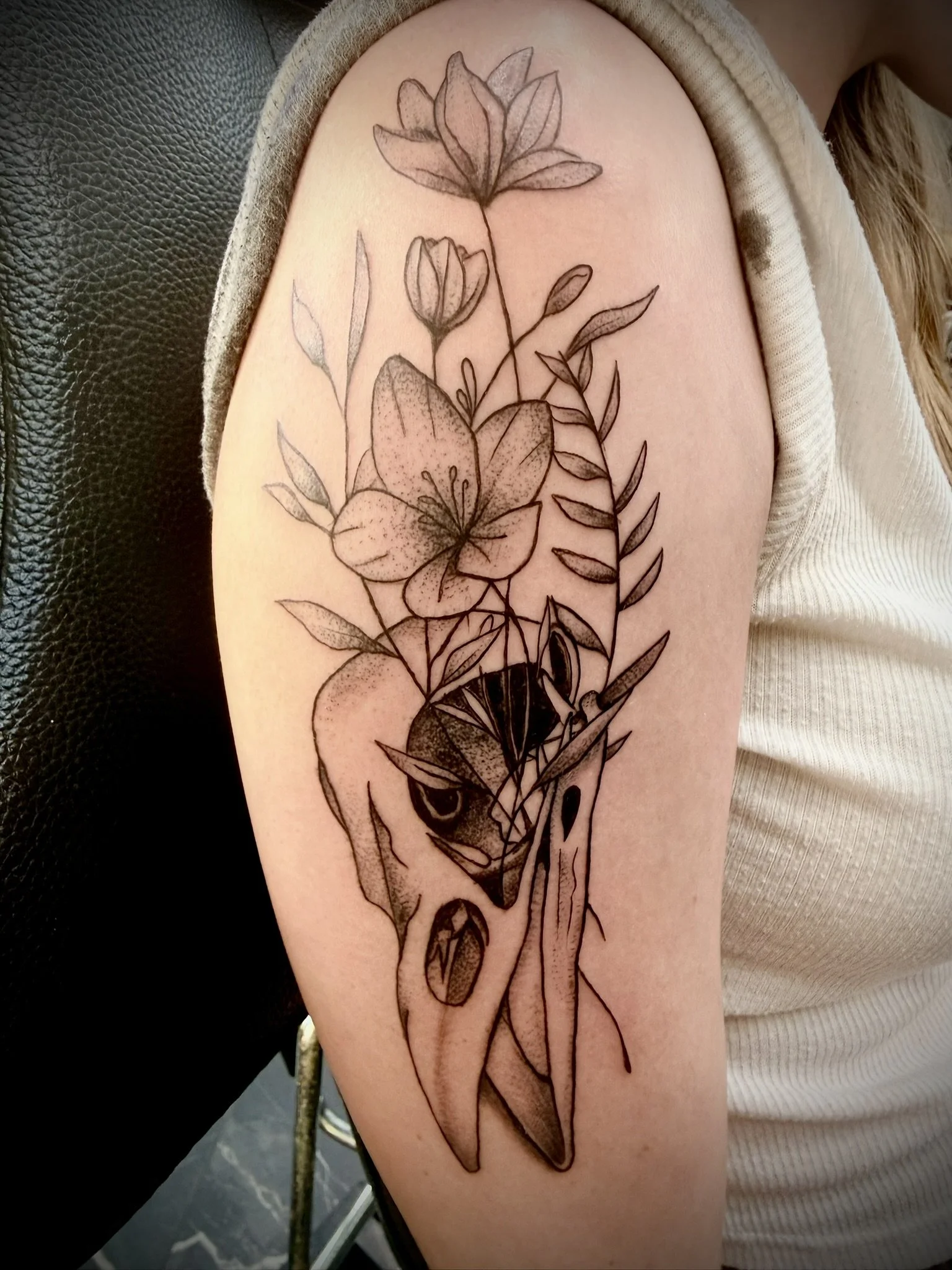 Black ink outline tattoo of two lilies on person's upper arm, with leaves and stems, partially covered by beige sleeve.
