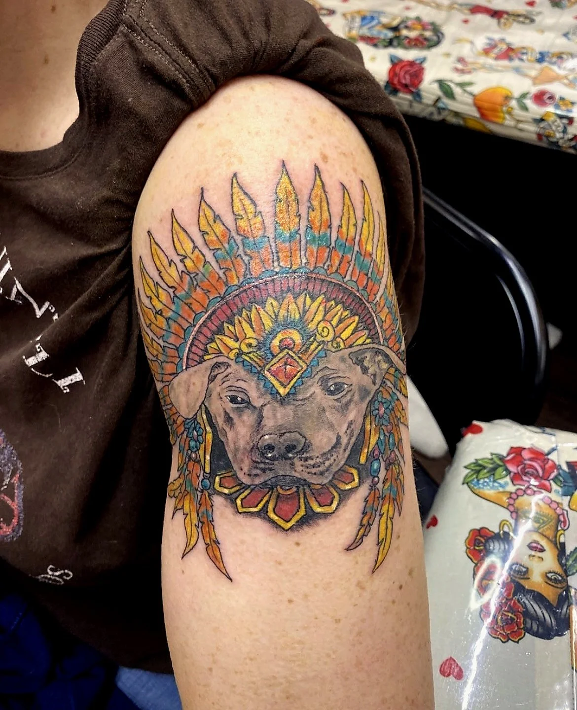 Tattoo of a dog wearing a colorful feathered headdress, reminiscent of Native American style, on a person's upper arm.