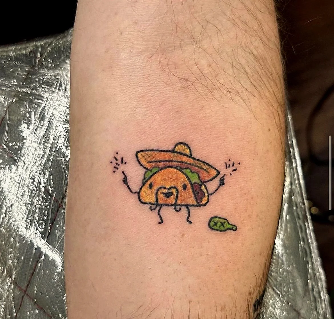 Cute tattoo of a taco with arms, legs, and a smiling face, wearing a sombrero, with a small green cactus nearby.