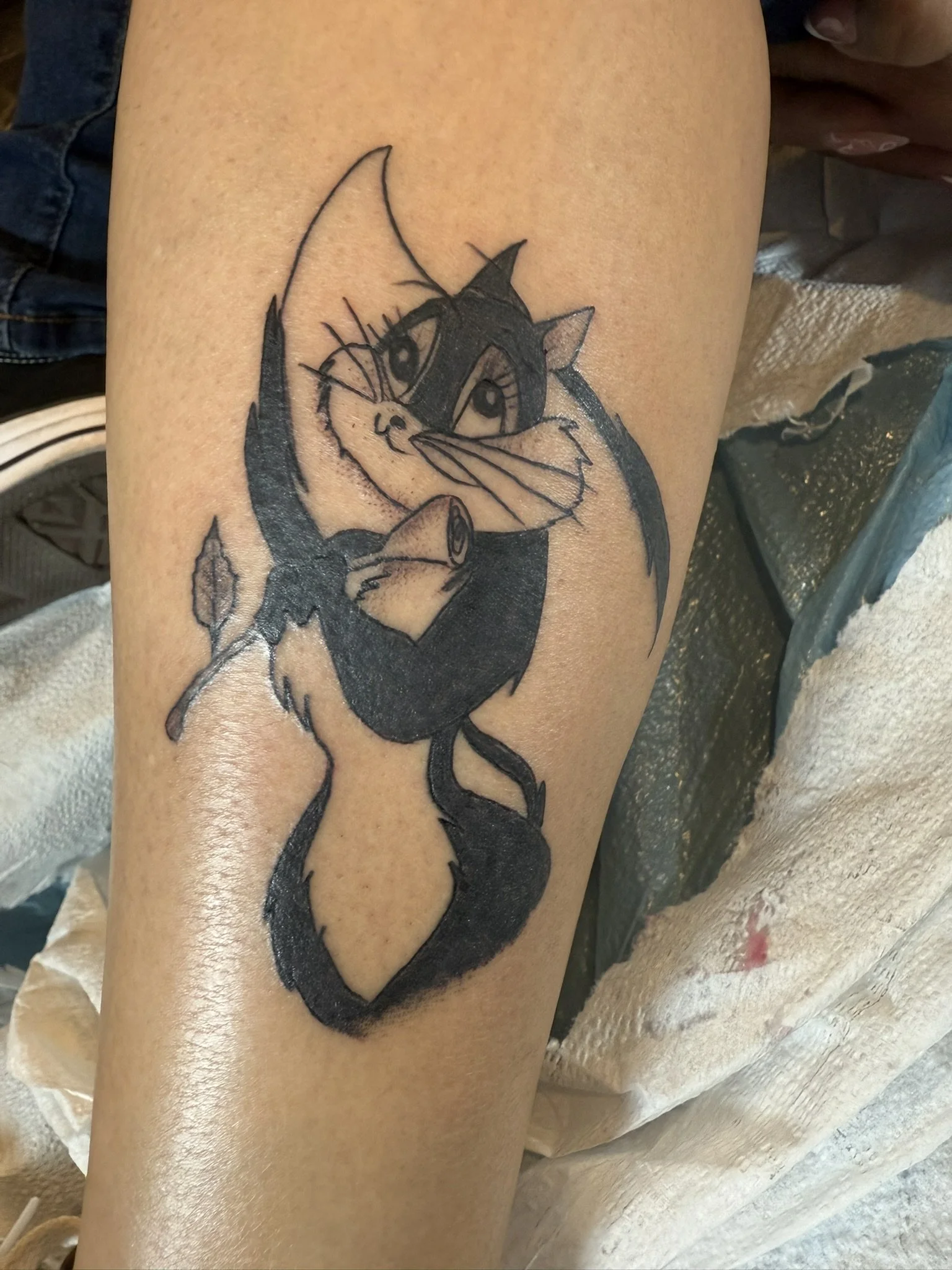 Black and gray tattoo of a cat with large eyes, sitting and holding its paws to its face, on a person's thigh.