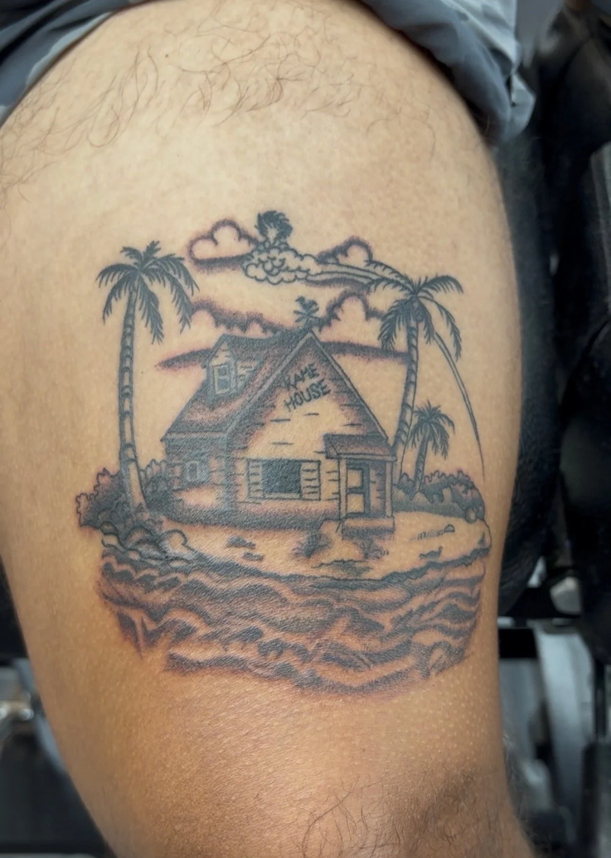 Tattoo of a house with a sign that says 'Beach House', surrounded by palm trees, clouds, and wavy water at the base.