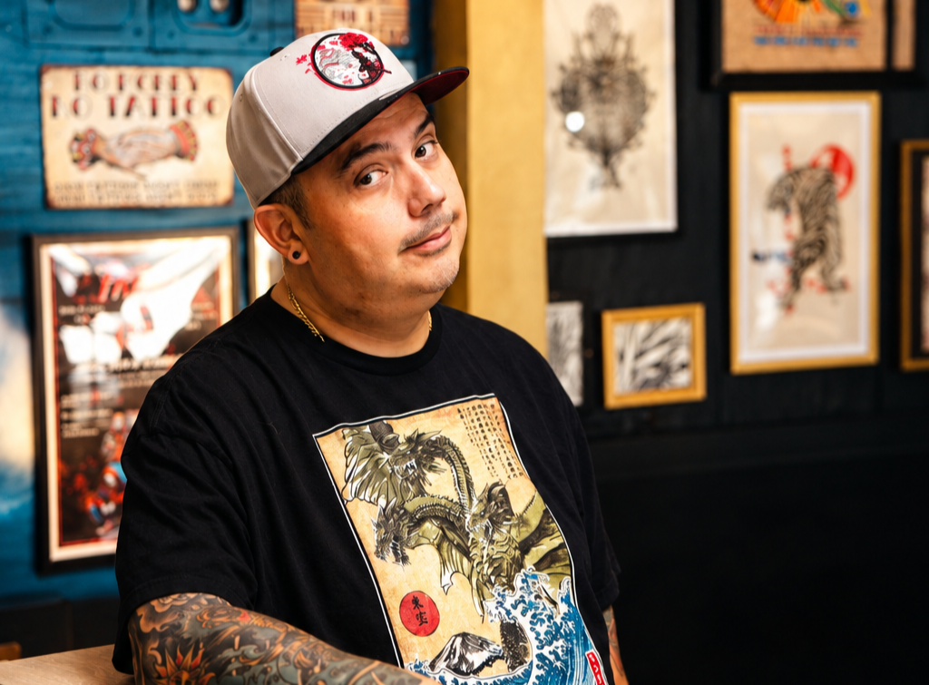 A man in a black T-shirt with Japanese-style art, wearing a beige and black baseball cap, stands in front of a wall decorated with various framed tattoo flash art and posters in a tattoo studio.