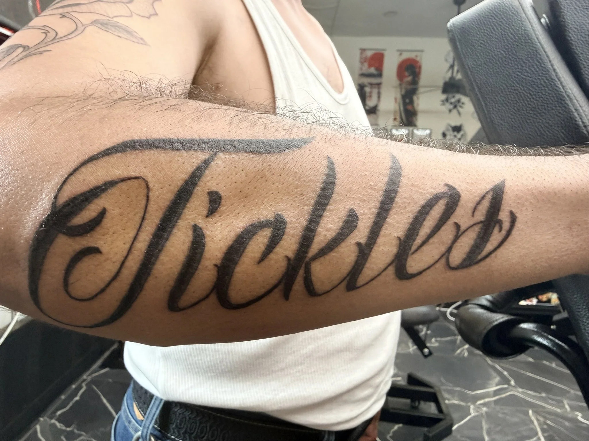 Close-up of a person's arm with a large tattoo in black ink that reads "Faith" in cursive script. The person is wearing a white sleeveless shirt, revealing a tattoo of a flower on the shoulder. The background shows a tattoo parlor with chairs and pos