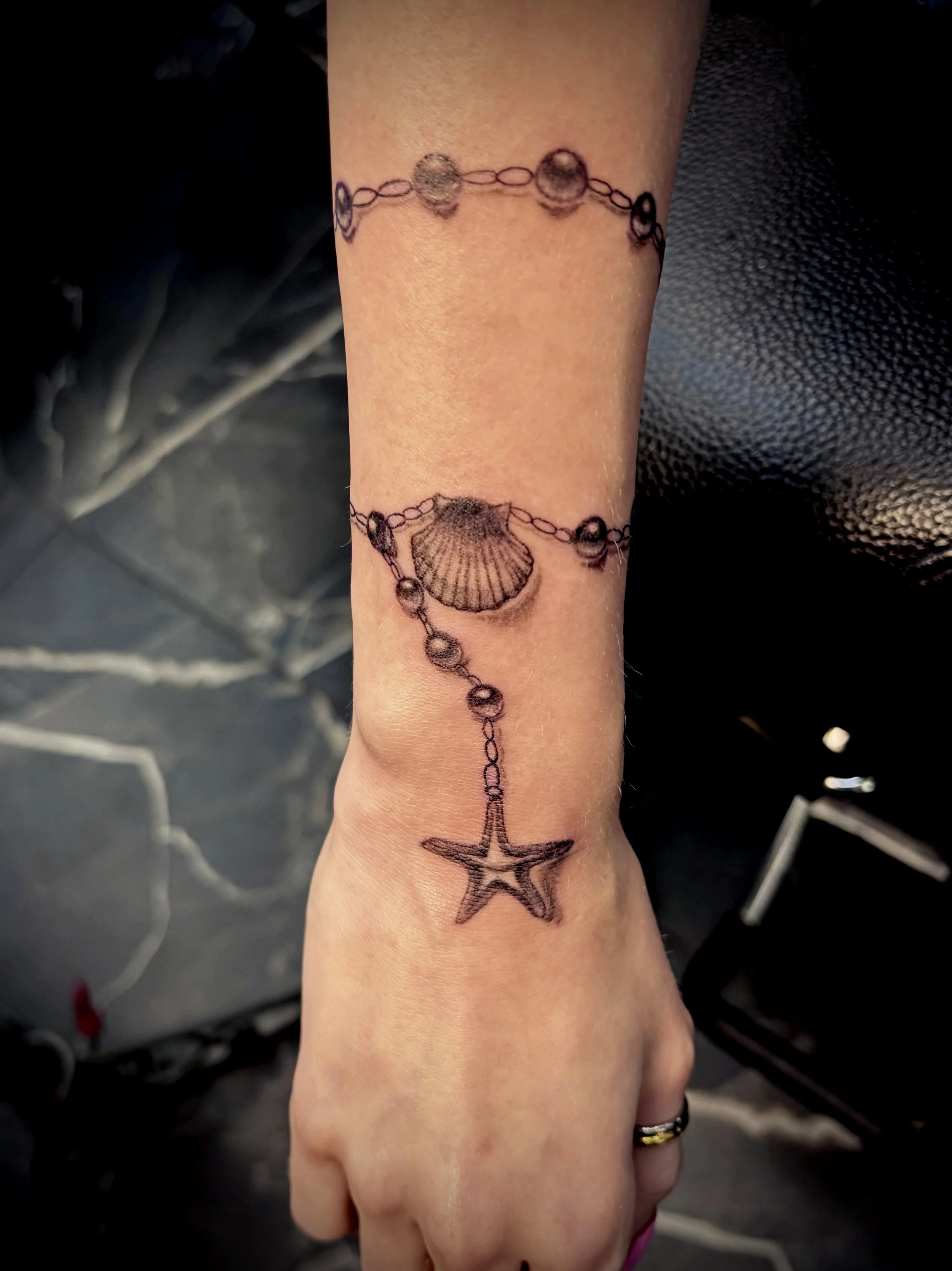 Tattoo of a seashell connected to a star charm by a chain of small beads on a person's wrist.