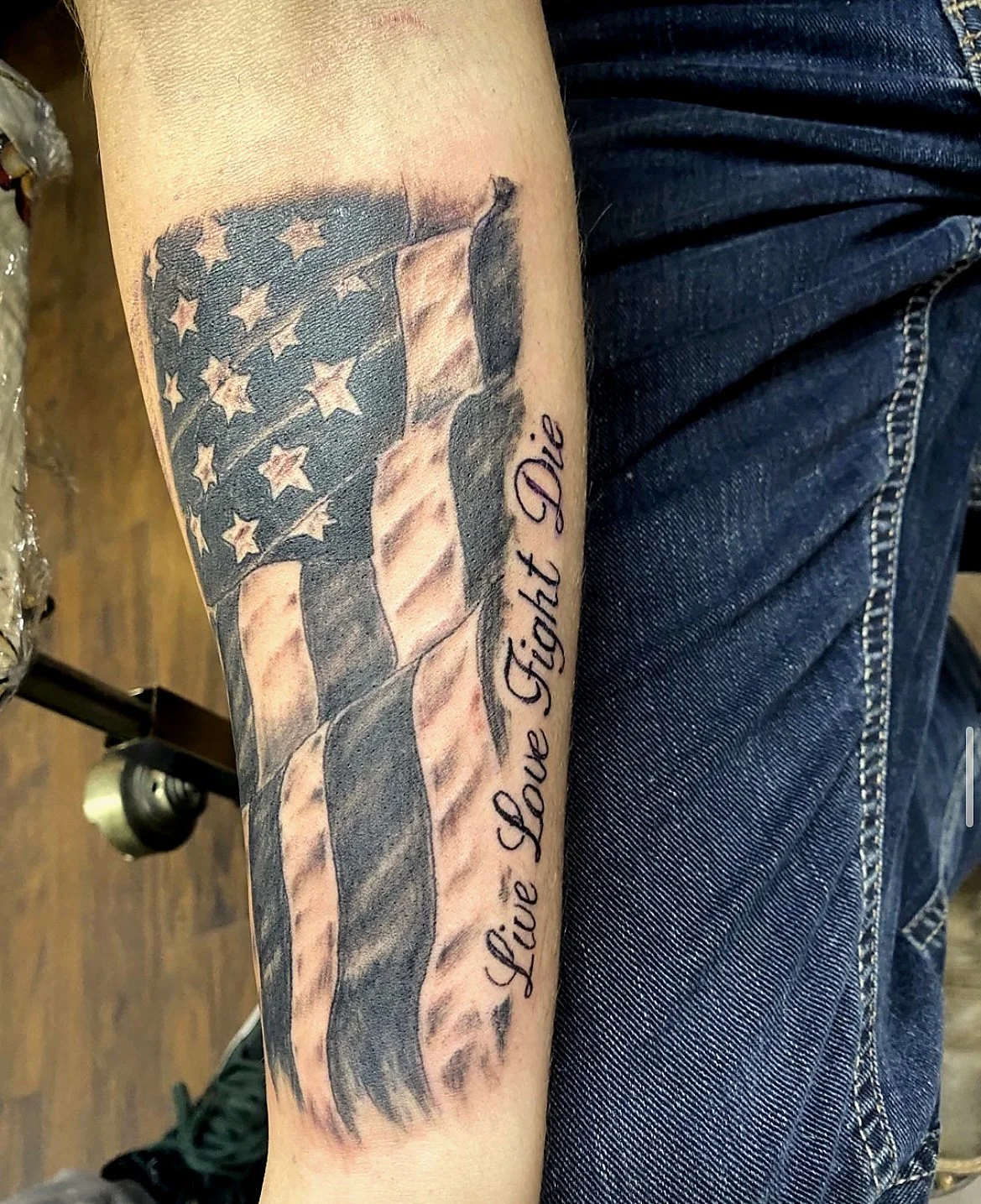 Tattoo of the American flag with a slogan 'Live, Love, Fight, Die' on a person's forearm.