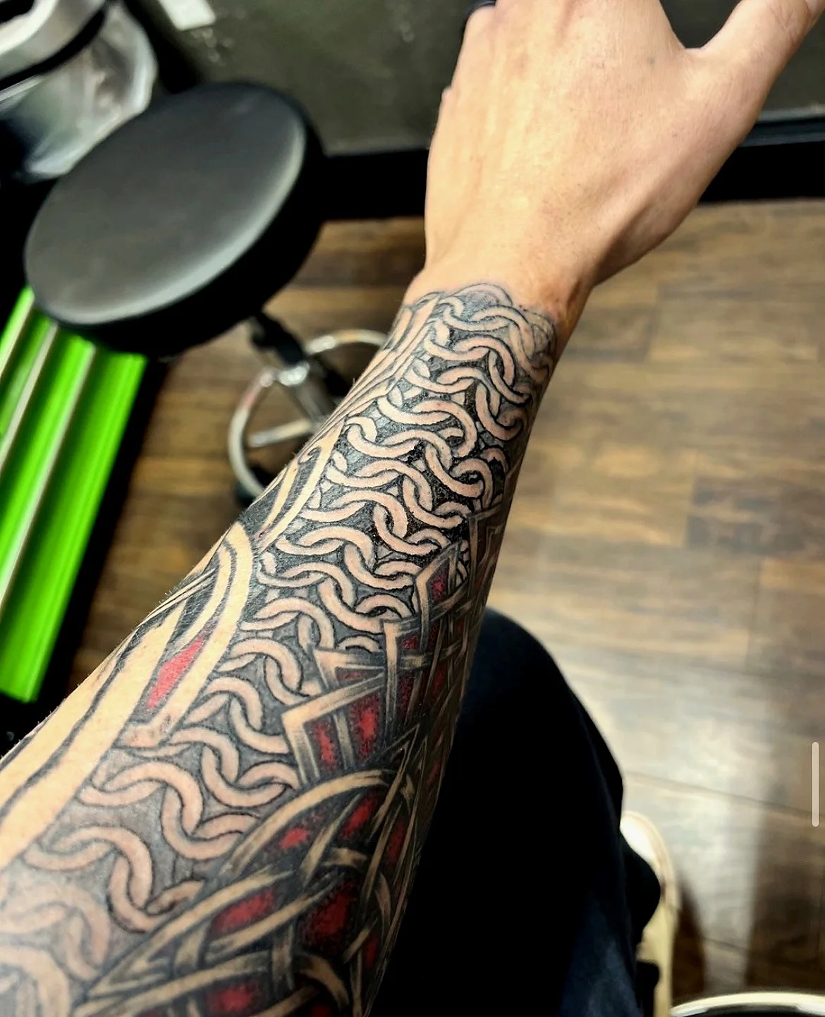 Close-up of a tattooed arm with interwoven chain-link pattern and red and black accents, extending from wrist to elbow, set against a brown wooden floor background.