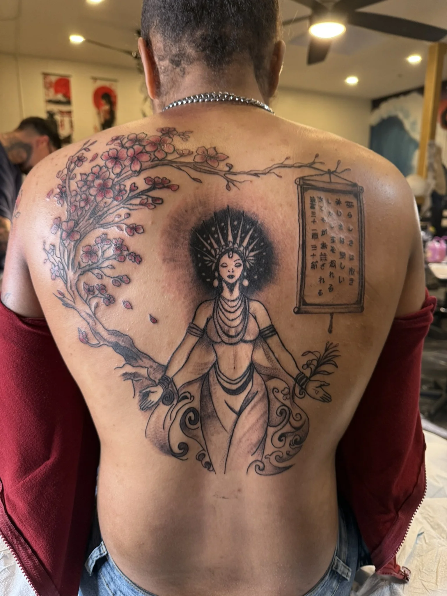 Tattoo on person's back featuring cherry blossom branch, a framed Japanese calligraphy scroll, and a stylized female figure with a radiant headdress, in black and red ink.