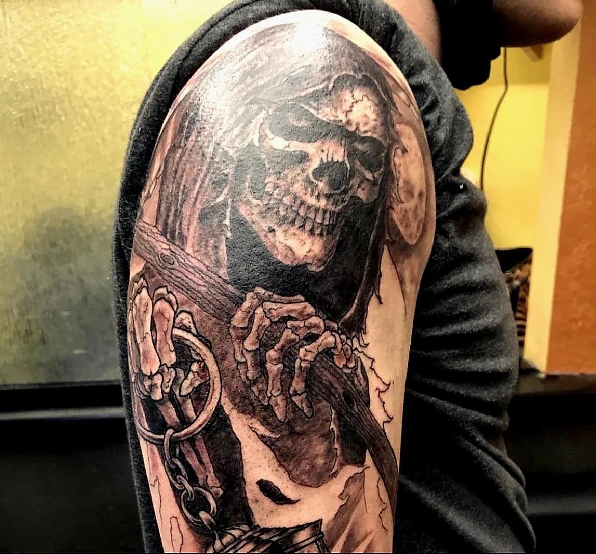 A detailed black and gray tattoo on a person's upper arm of a skeleton with a skull, holding a chain and a sword.