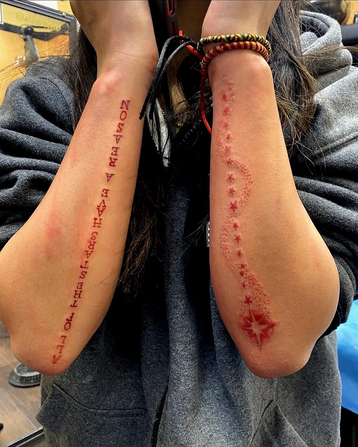 Person with two tattoos on forearms; the right forearm has a red printed text tattoo, and the left forearm has a red star and dot constellation tattoo along with bracelets.