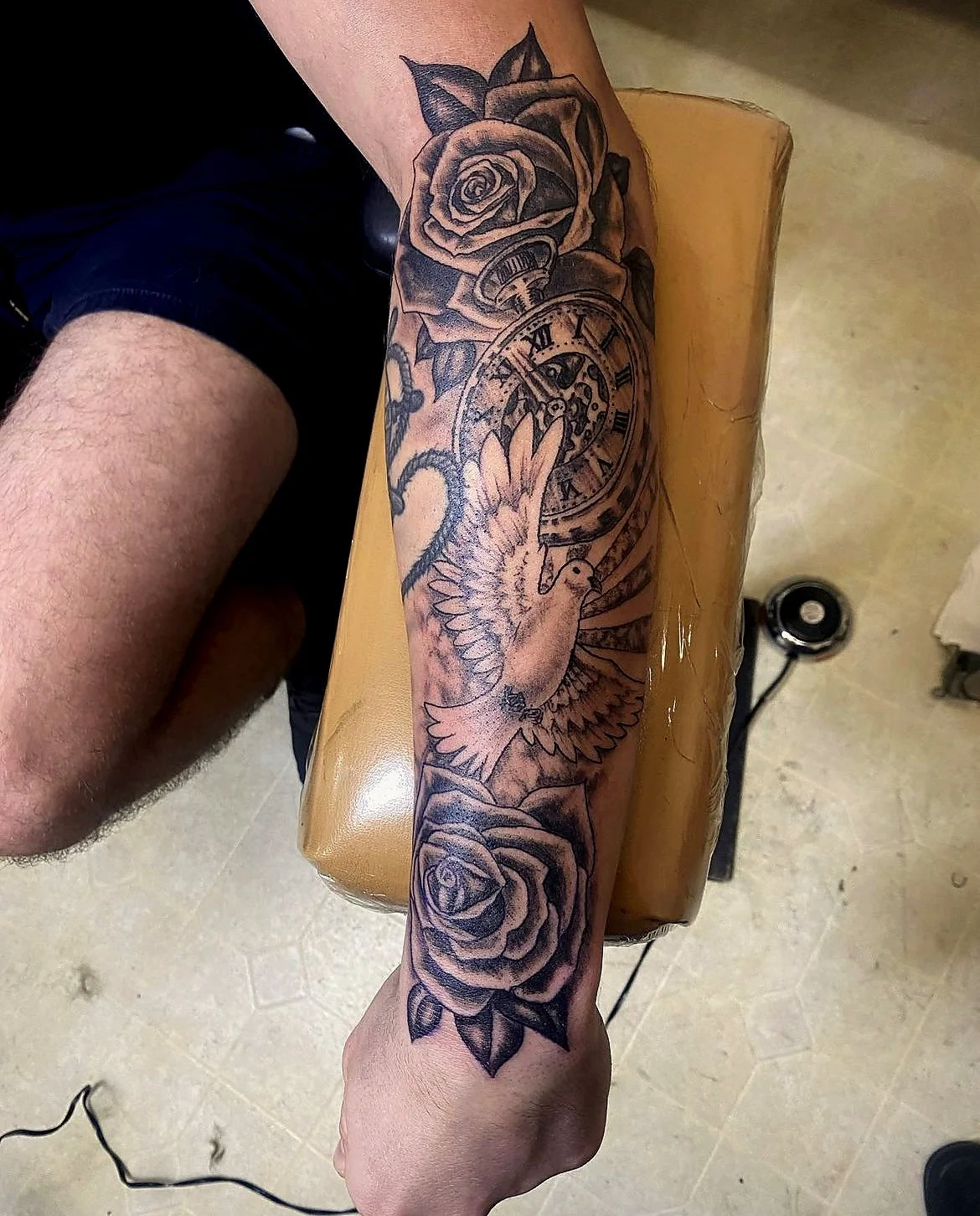 Tattoo of roses, a pocket watch, a dove with outstretched wings, and sunburst rays on a person's forearm.