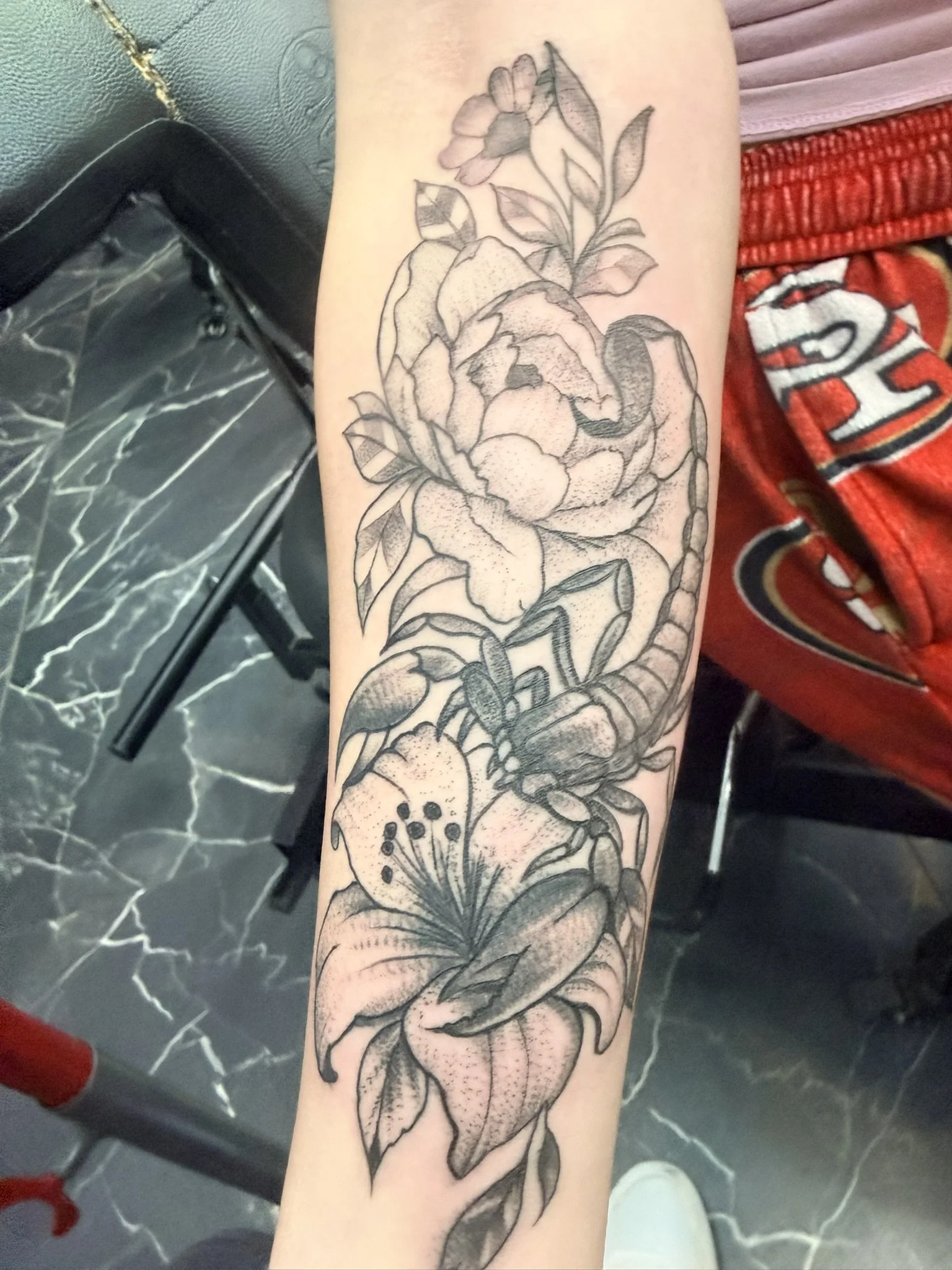 A black and gray tattoo of a large flower, possibly a lily or a peony, with detailed petals, leaves, and stems on a person's forearm.