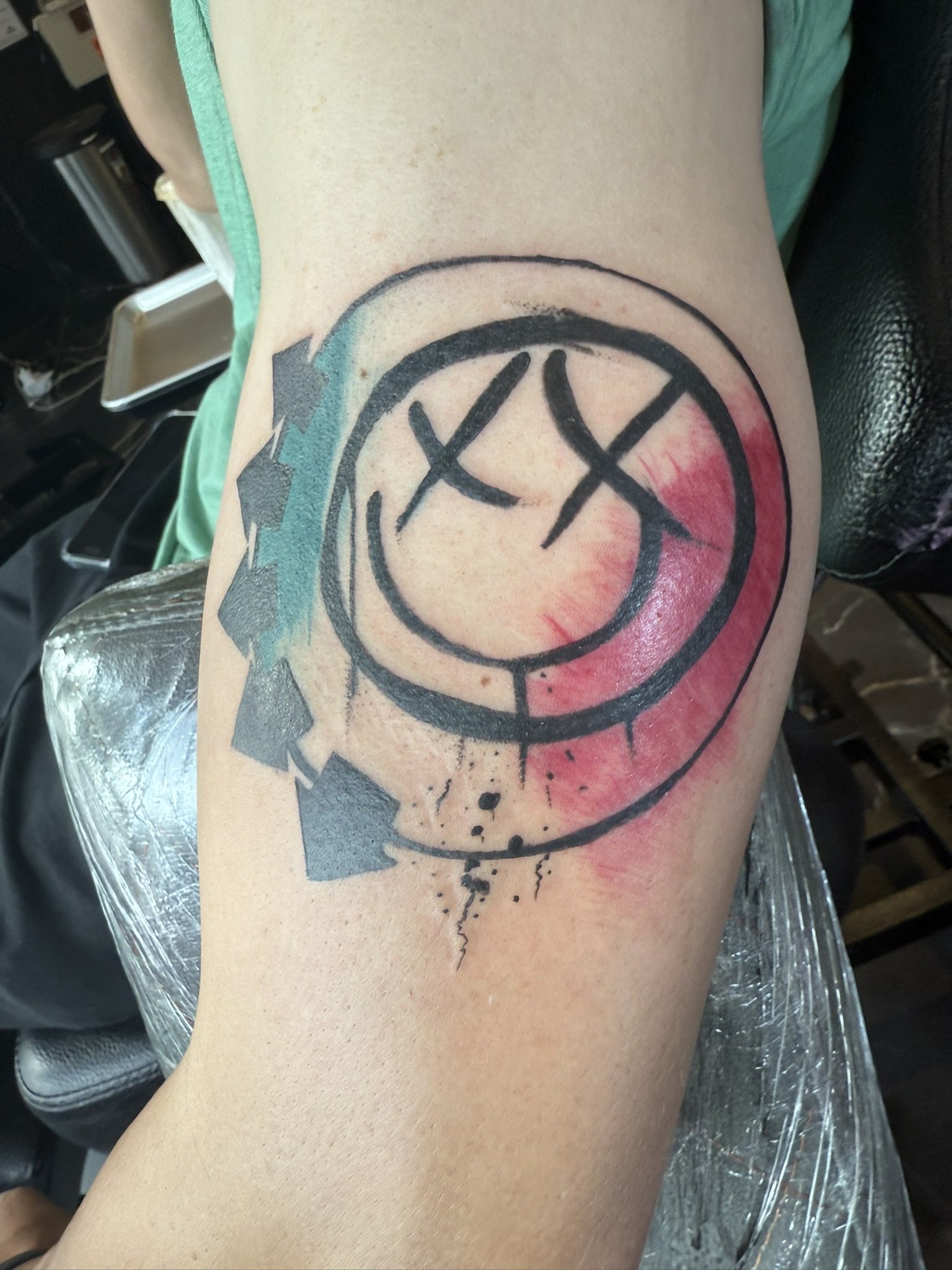A tattoo of a clock with roman numerals and abstract coloring on a person's upper arm.