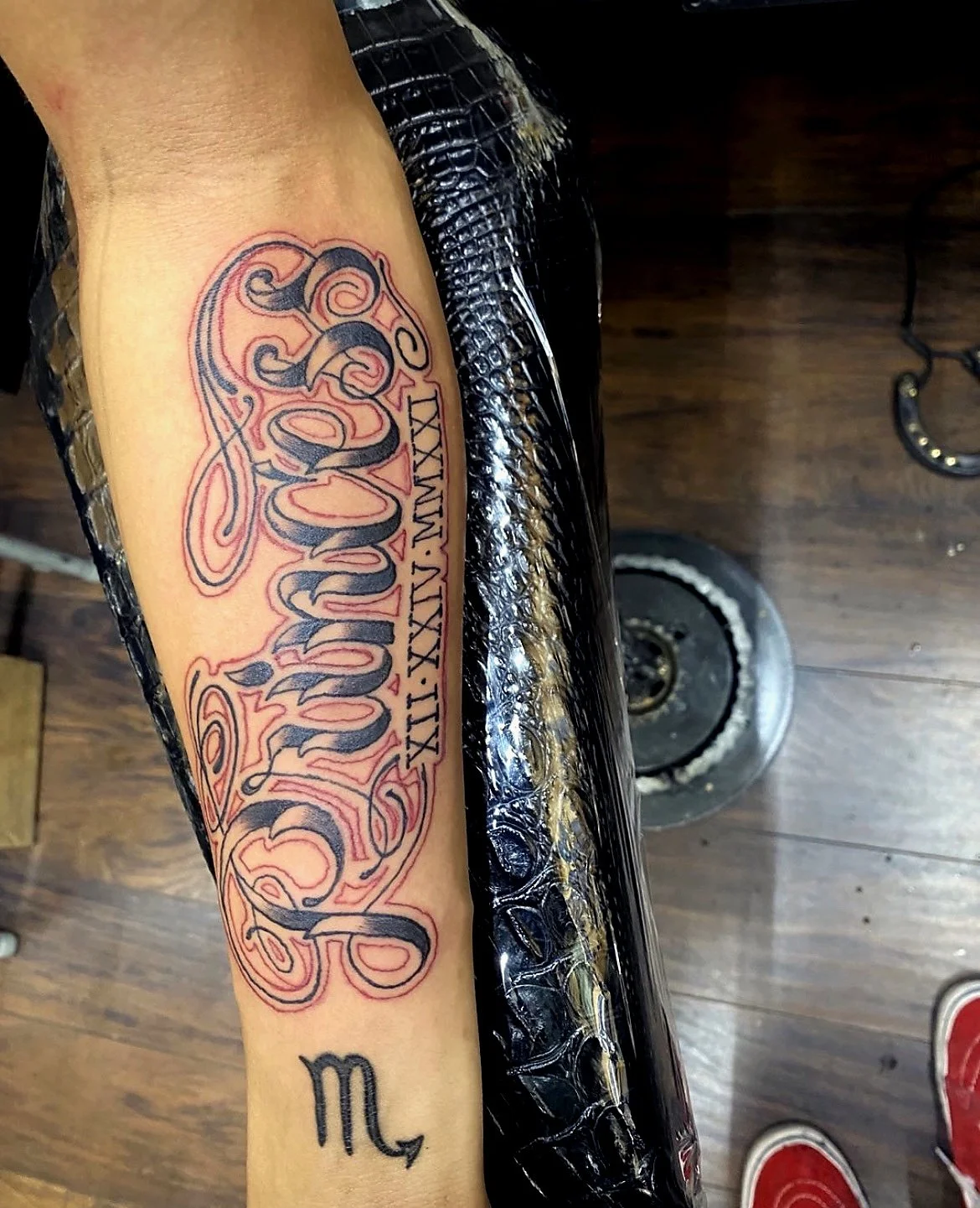 A tattoo on a person's forearm with ornate black script and Roman numerals, with a horseshoe visible in the background on a dark wood floor.