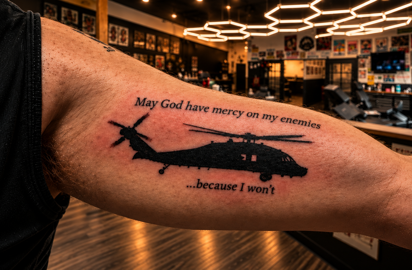 Tattoo of a helicopter accompanied by the quote, 'May God have mercy on my enemies...because I won't,' on a person's upper arm in a tattoo parlor.