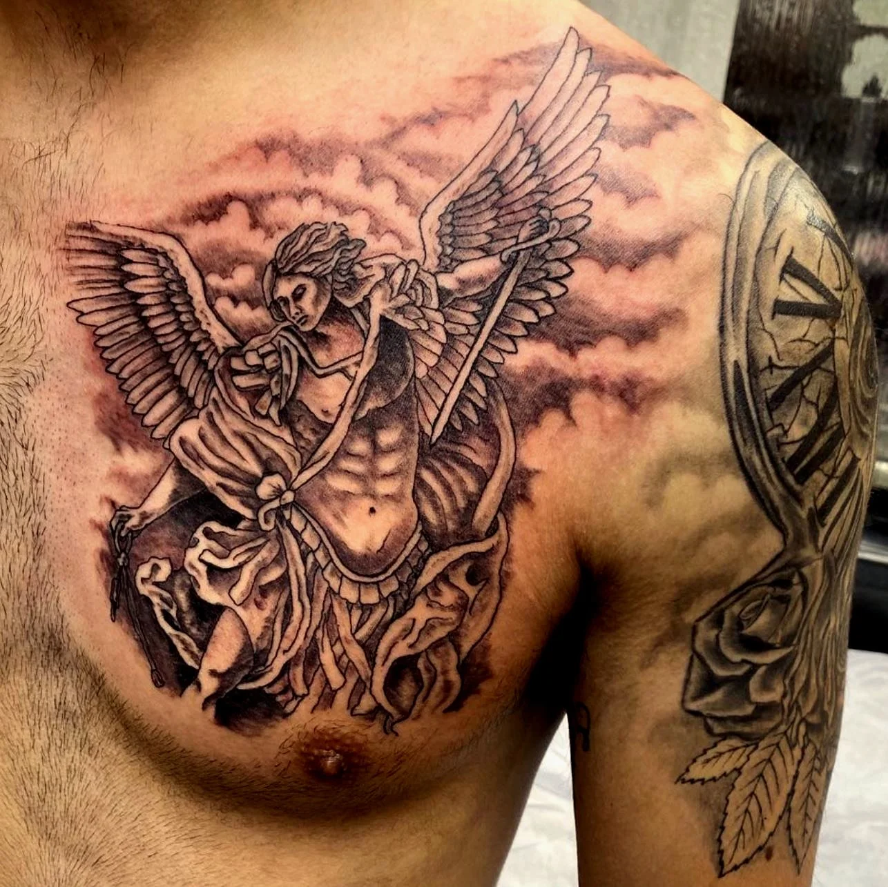 Black and gray tattoo of an angel with wings holding a staff, with clouds and a clock on the shoulder.