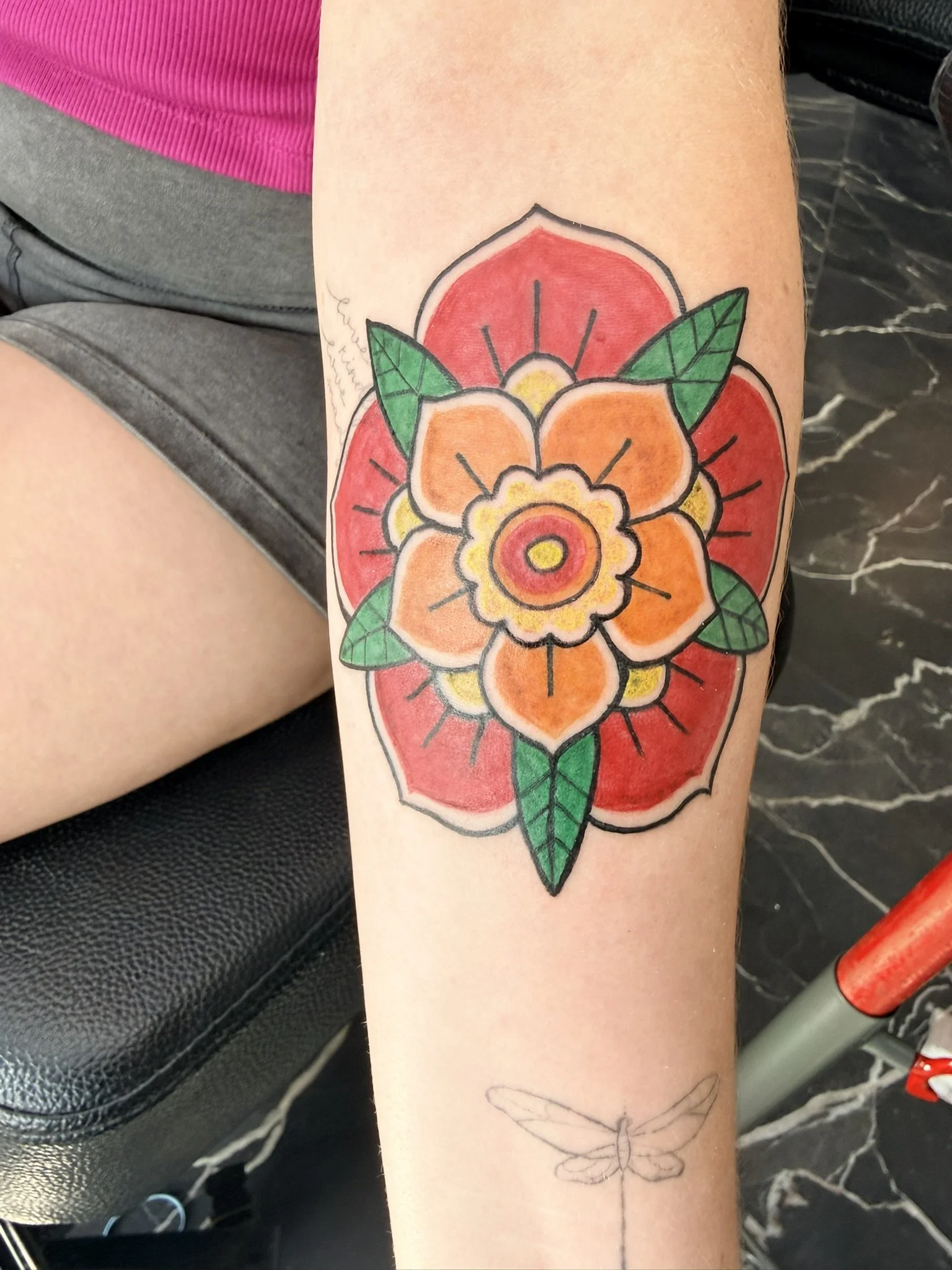 Colorful tattoo of a large flower with orange, yellow, and red petals, green leaves, and black outlining on a person's arm, with a outline of a dragonfly tattoo below.