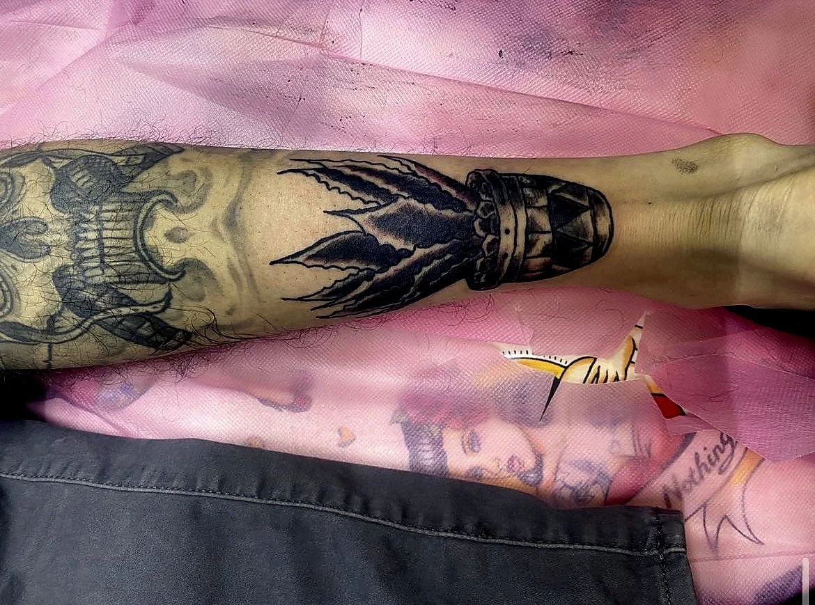 Tattooed arm lying on pink surface with additional tattoos visible on another person's hand nearby.