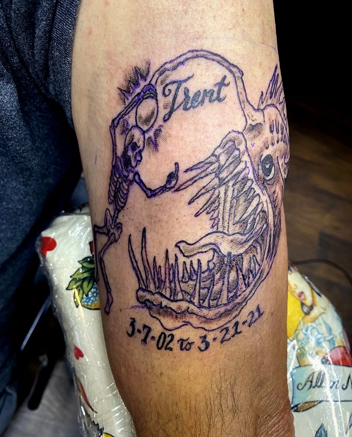 Tattoo of a dinosaur skeleton with the word 'Trust' inside the skull, and dates 3-7-02 to 3-22-21 below the skull.