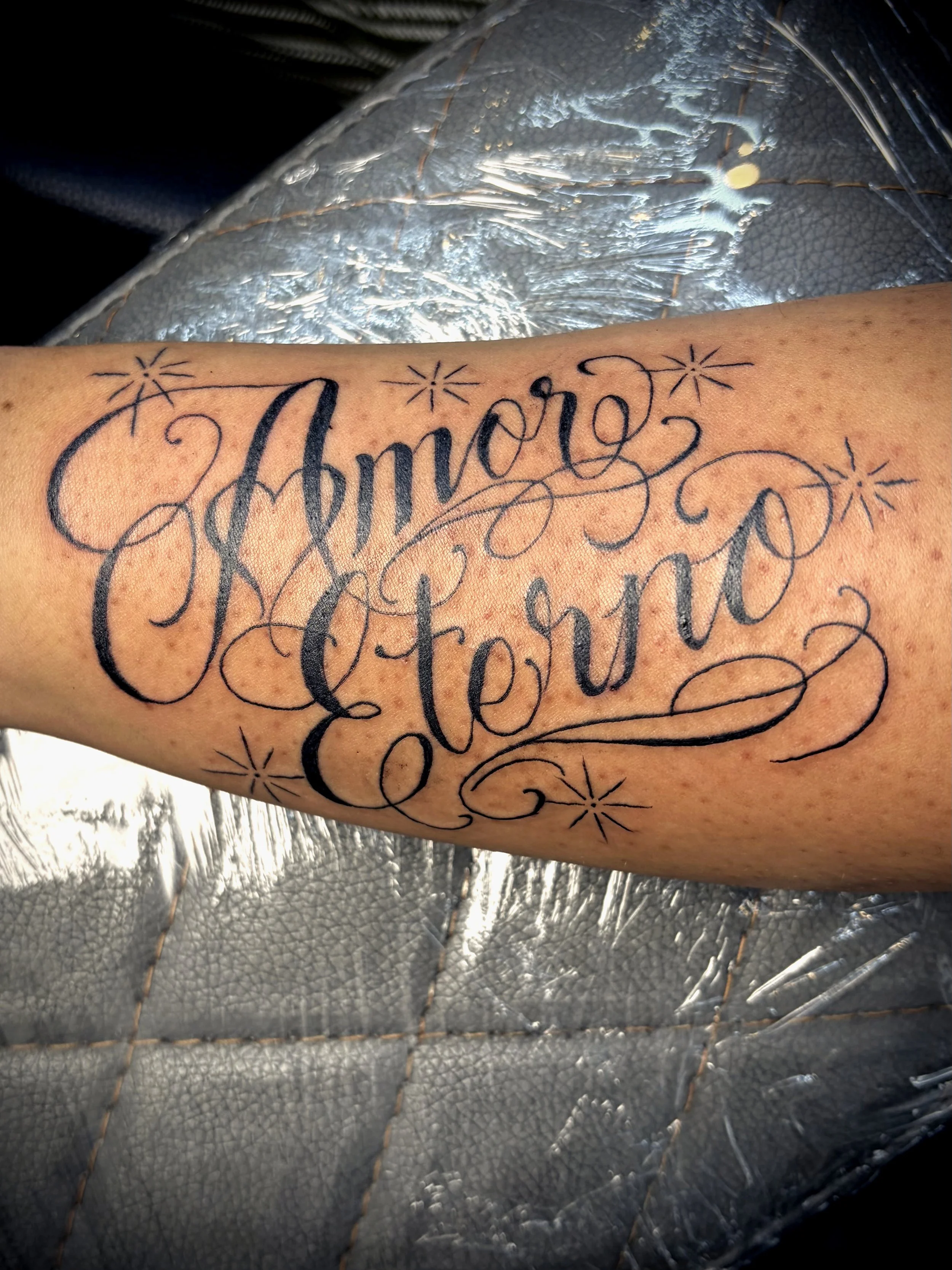 Close-up of a human arm with a tattoo that reads 'Ambitious' in cursive script, surrounded by small decorative stars, resting on a silver textured surface.