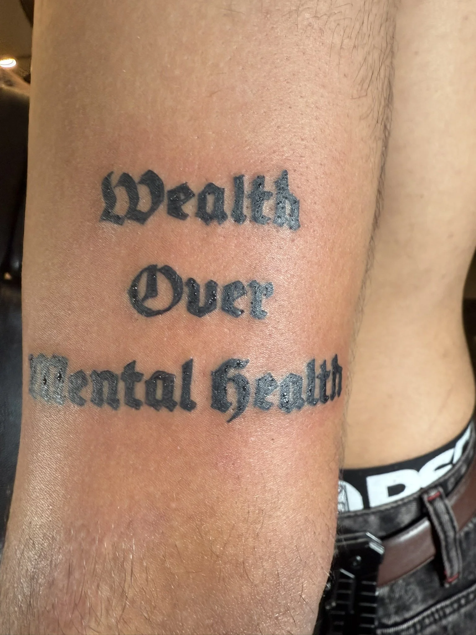 A close-up of a person's upper arm with a tattoo that reads 'wealth Queen' written in black ink in a Gothic font style.