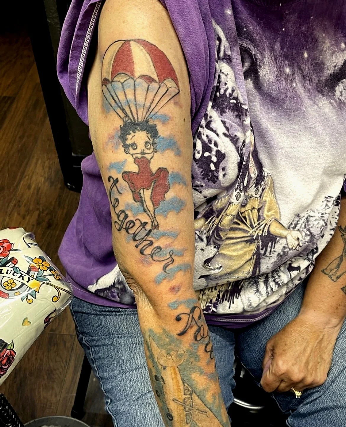 Tattoo of Betty Boop holding an umbrella with clouds and the word 'together' on a person's upper arm