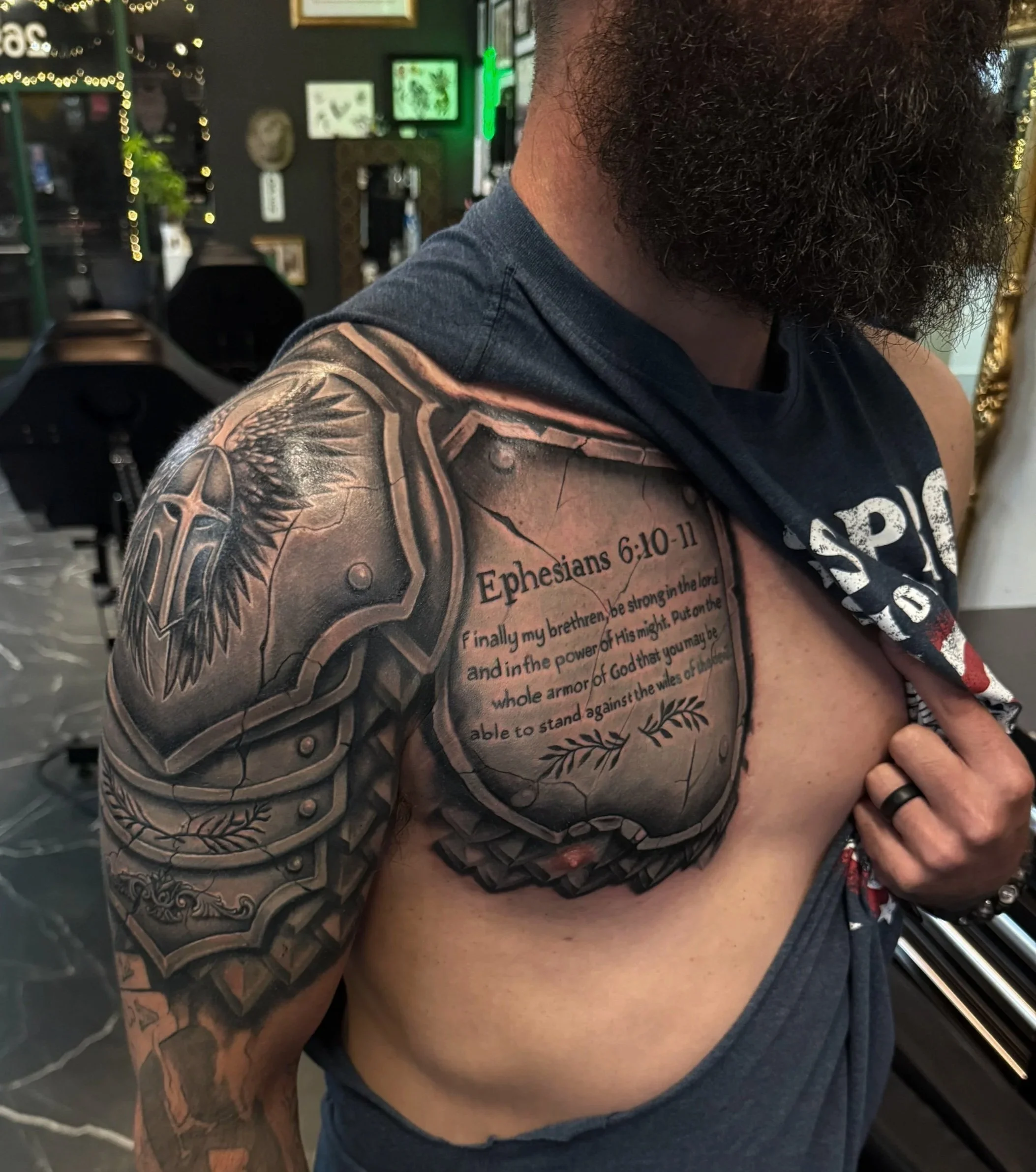 A shirtless man with a full beard, wearing a black sleeveless shirt and a black ring, showing a detailed tattoo sleeve on his left arm with religious text and imagery, in an interior setting with decorated walls and furniture.