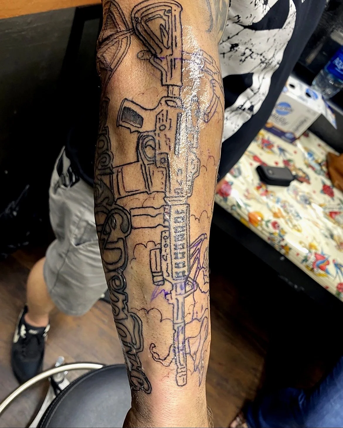A tattoo on a person's forearm featuring a detailed sketch of a firearm with clouds and smoke around it.