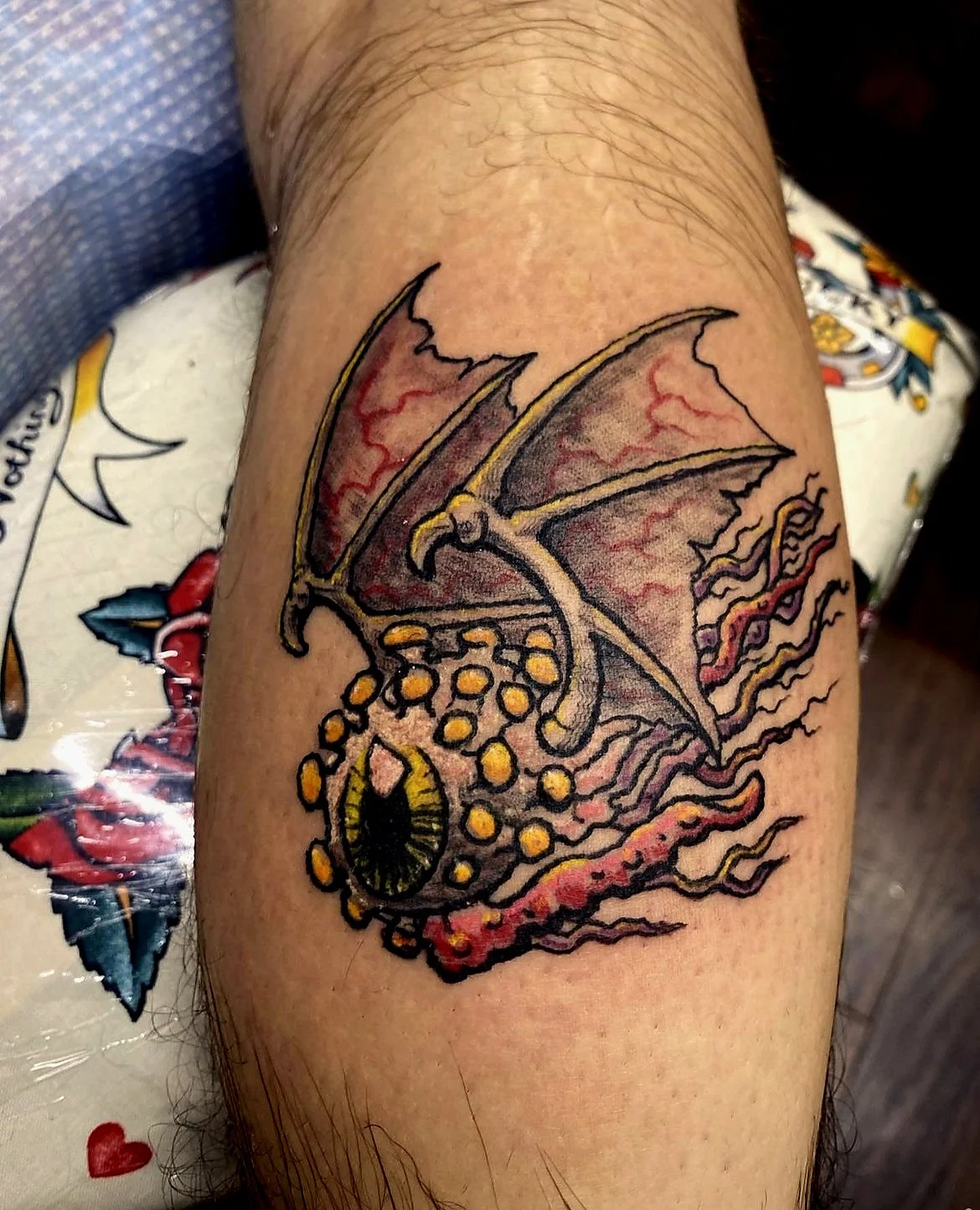 Tattoo of a dragon with red and yellow wings, a circle with an eye inside, and surrounded by flames on a person's arm.