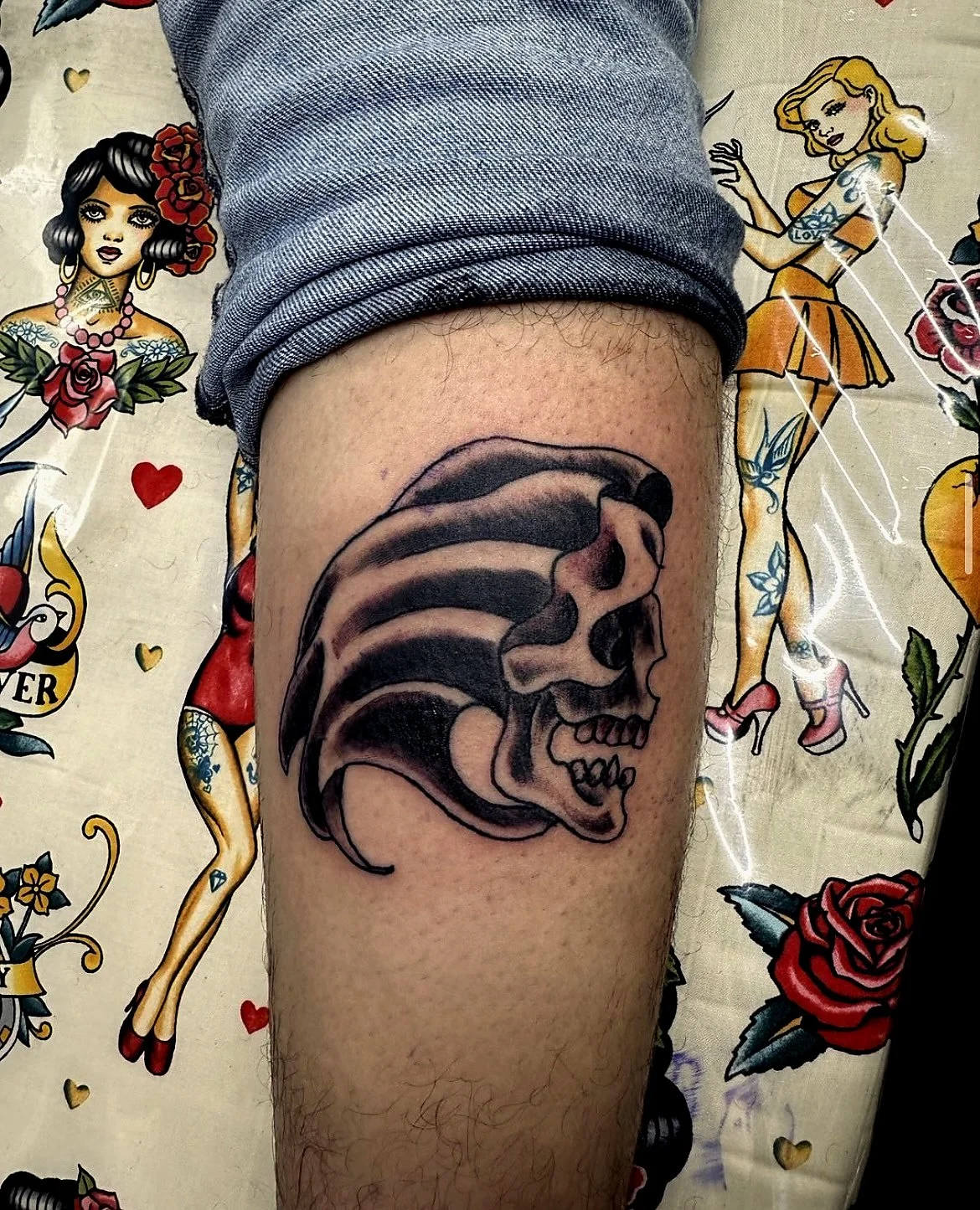 Tattoo of a human skull wearing a mohawk on a person's thigh, with a background featuring tattoo-style pin-up girls and hearts.
