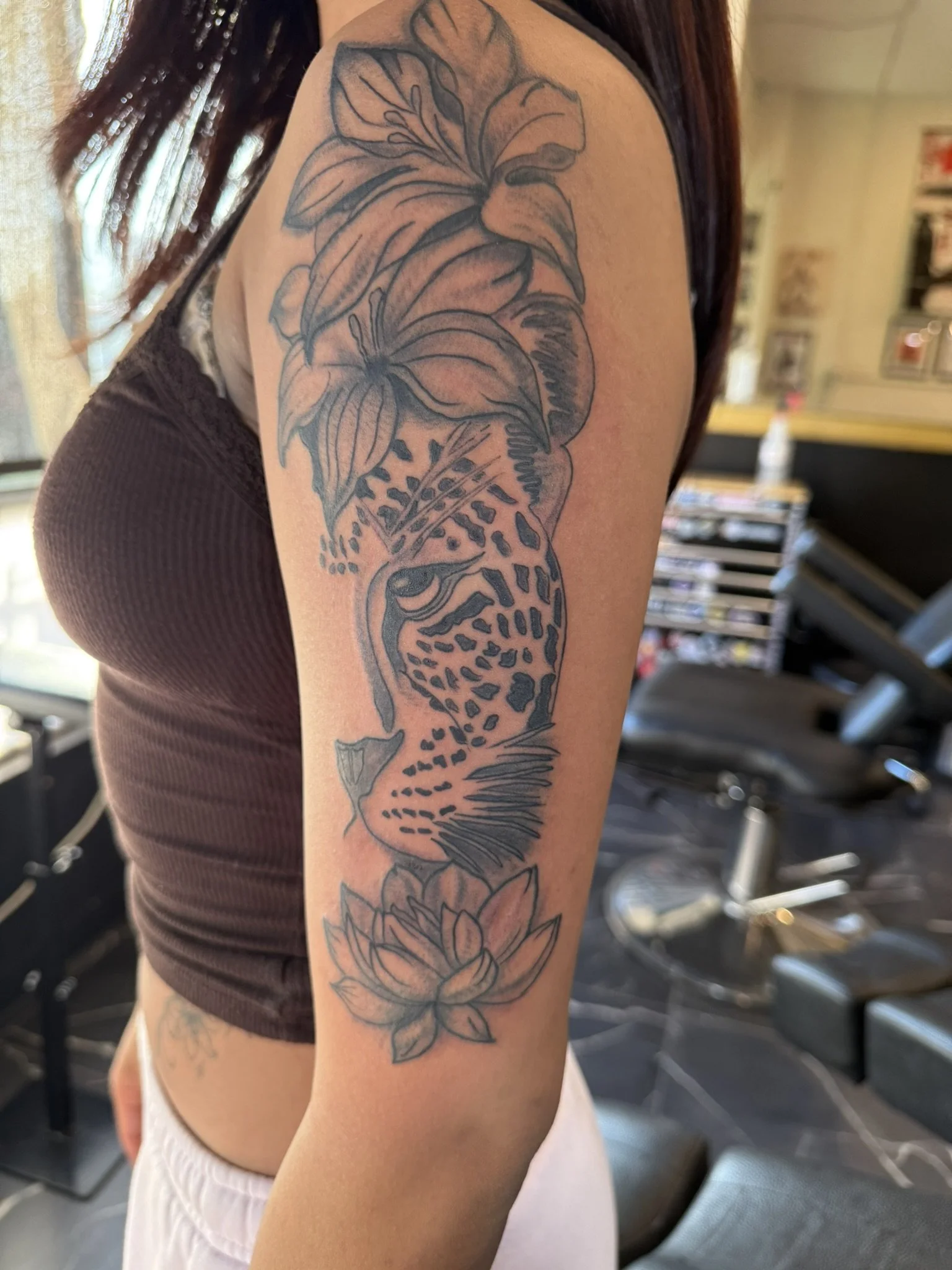 A person's upper arm and shoulder with a detailed tattoo of a fish with floral elements on the arm, in a tattoo studio with chairs and shelves in the background.