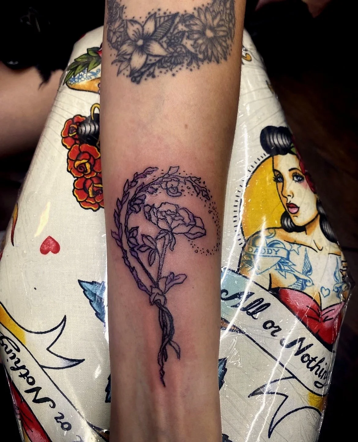 A person's forearm with a tattoo outline of a rose and leaves, surrounded by dotted lines. In the background, there are other tattoos and a colorful sheet with vintage-style artwork and text.