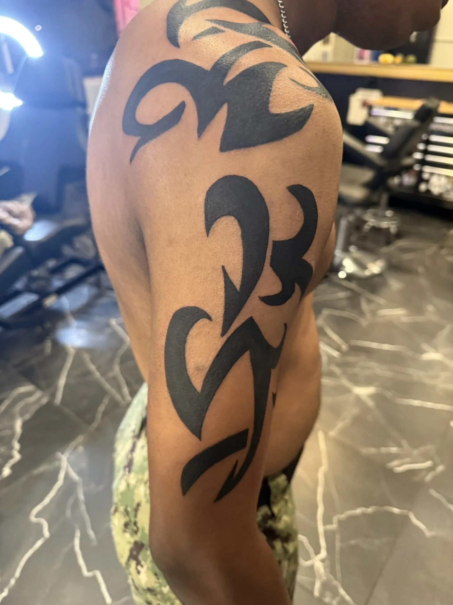 A person's upper arm and shoulder with a large, black tribal tattoo design. The person is indoors, with a blurred background including a chair and some objects.