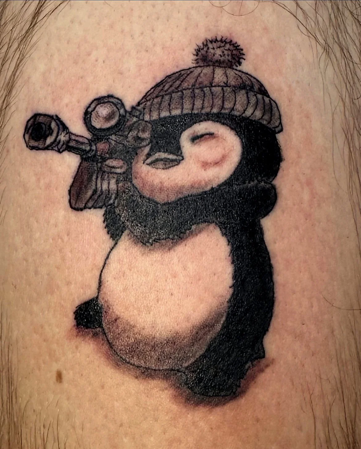 Tattoo of a penguin wearing a knit hat and holding a camera, with a pink heart-shaped object on its chest.