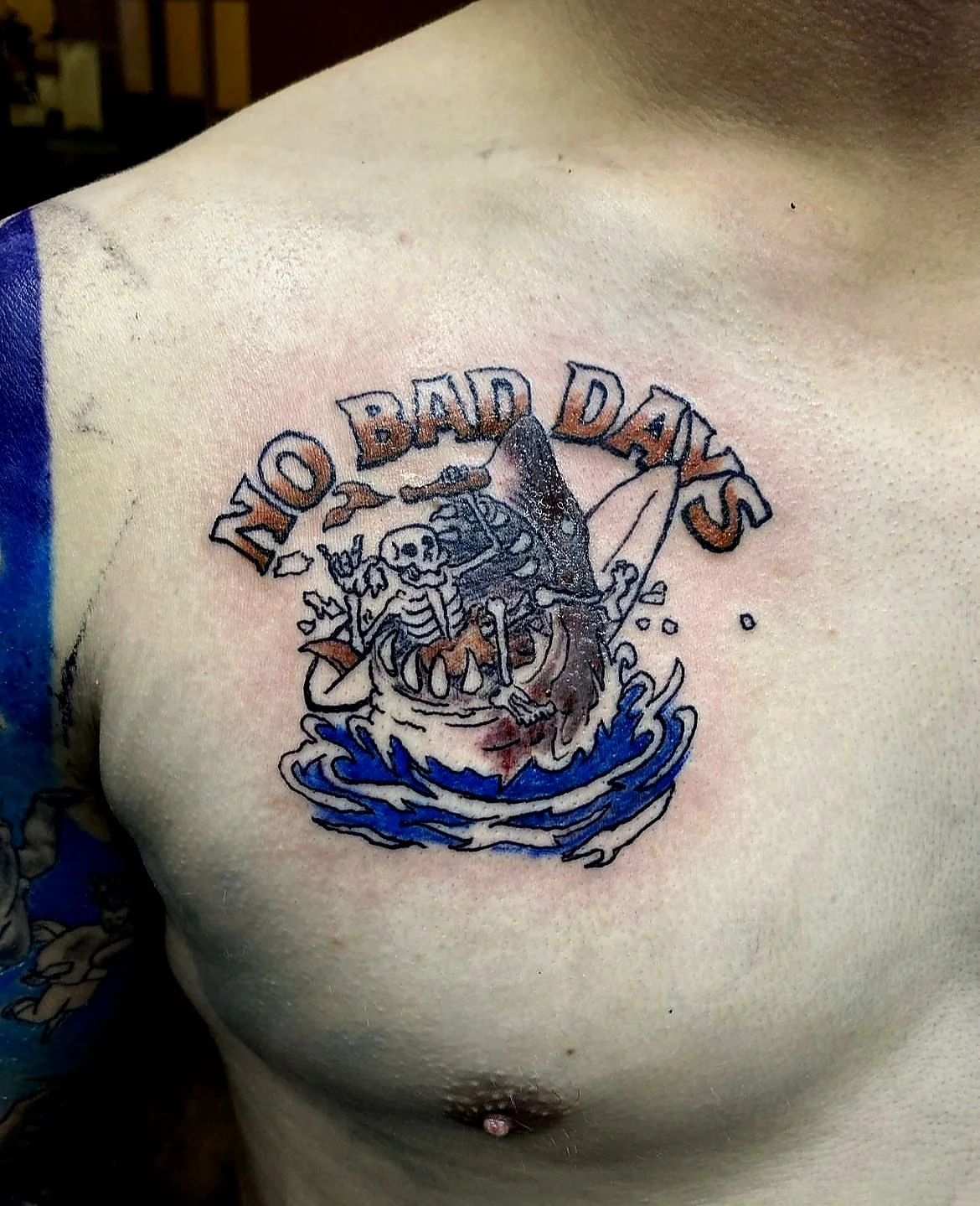 Tattoo on person's chest showing a skeleton in a boat with a shark fin, surrounded by water splashes. Above the boat, the text reads 'NO BAD DAYS' in bold letters.