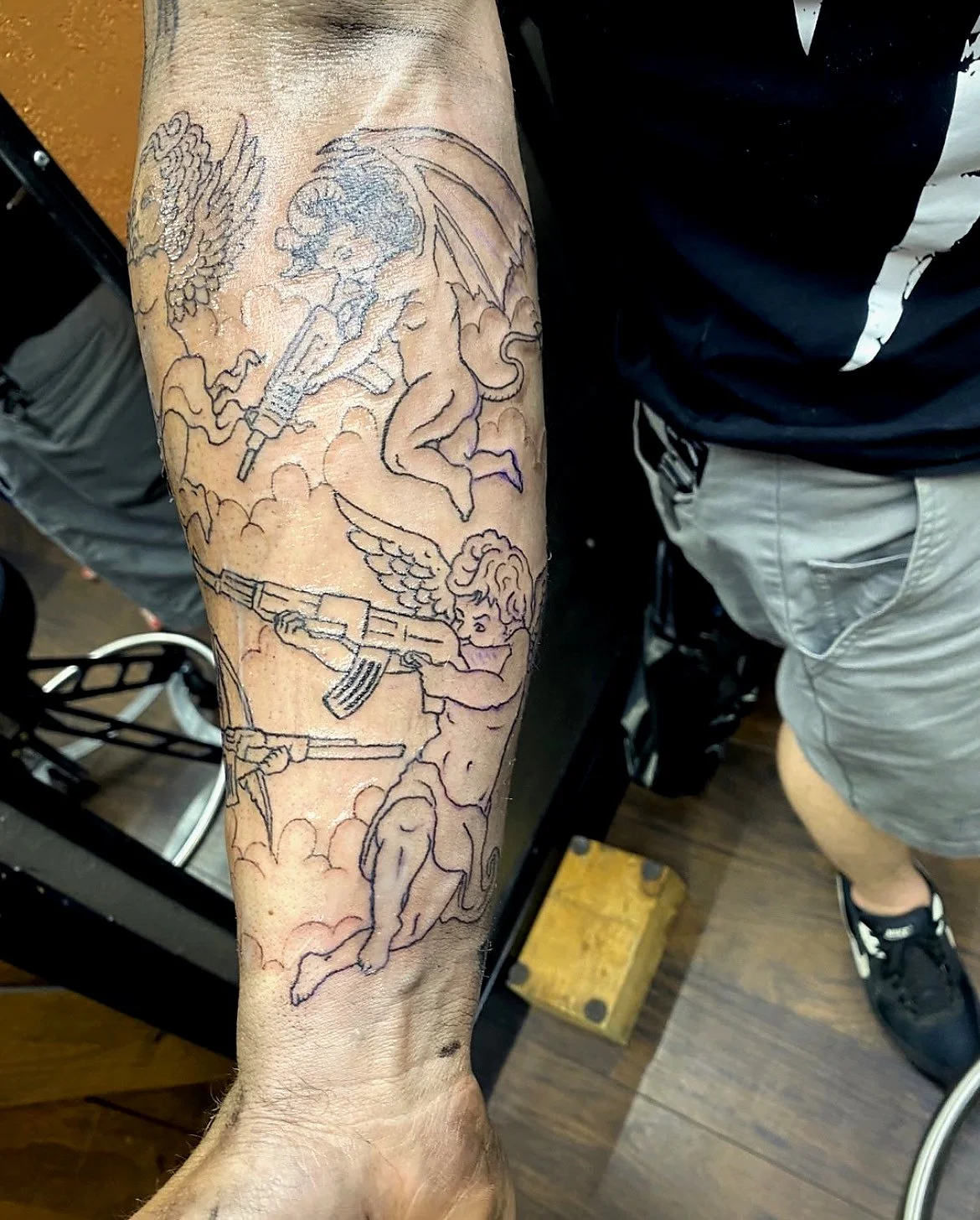 Tattoo of angels with wings, holding guns, surrounded by clouds, on a person's forearm.
