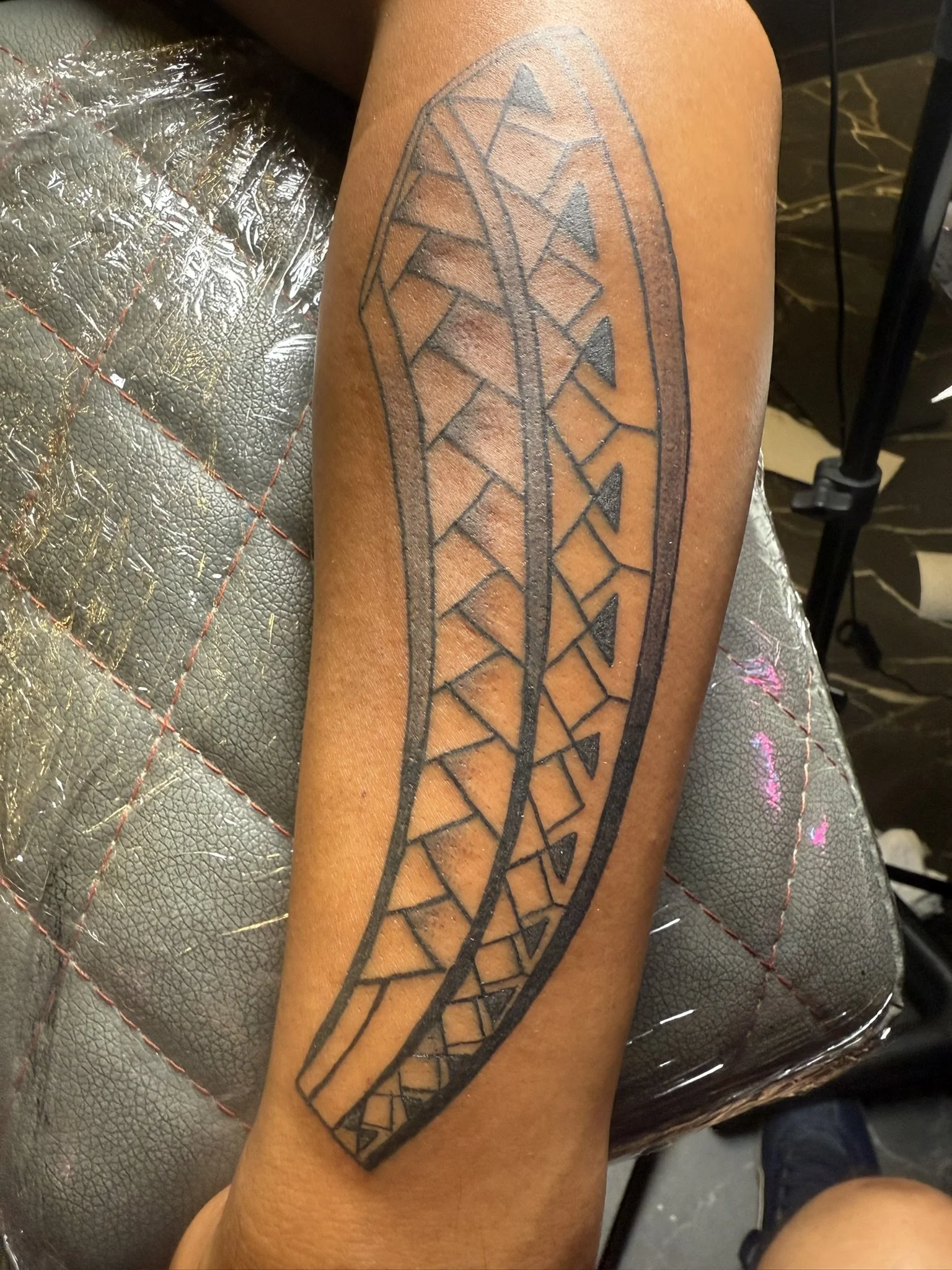 A black tribal-style tattoo on a person's forearm, featuring geometric shapes within an elongated oval outline.
