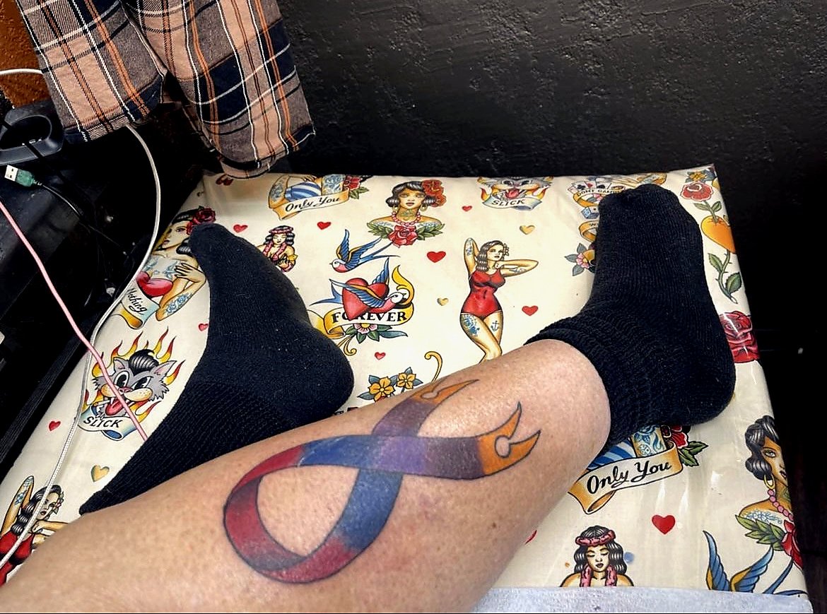 A person's leg with a colorful tattoo of a bow on their calf, wearing black socks, resting on a tattoo-themed bedspread with vintage tattoo flash designs.