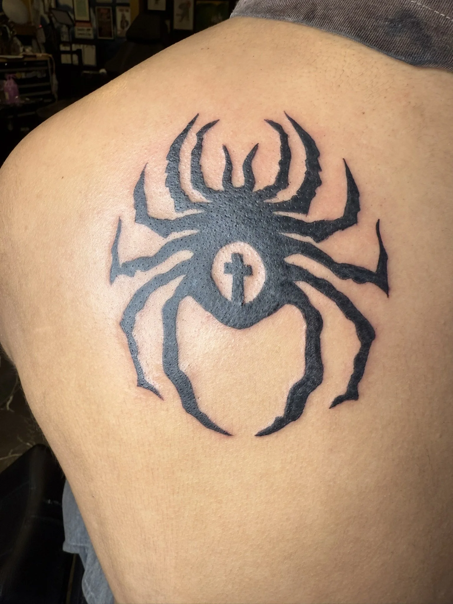 A black tribal-style tattoo of a spider with a cross in the center is on a person's shoulder and upper arm.